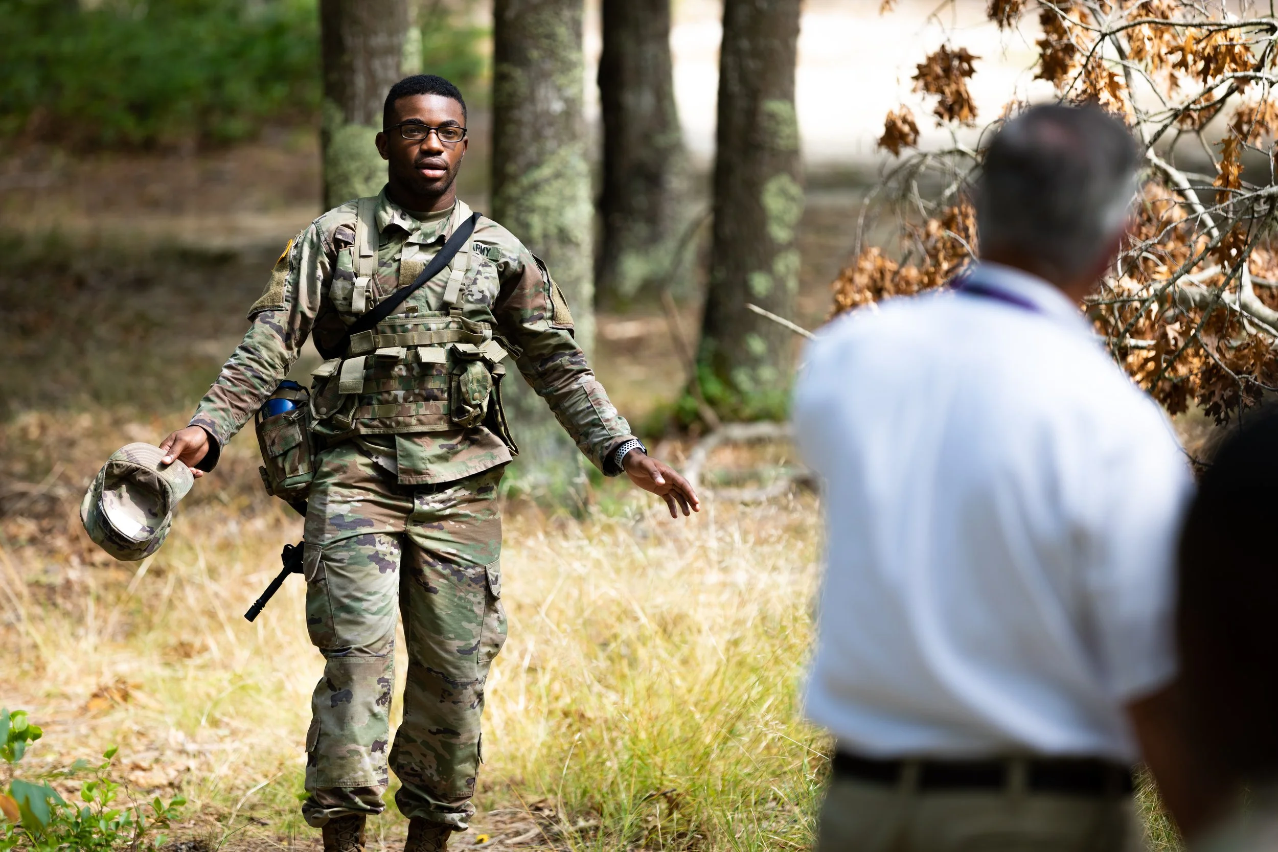 A soldier in camouflage uniform holding a helmet stands in a forested area. Another person, seen from behind, is wearing a white shirt and appears to be sitting or crouching.