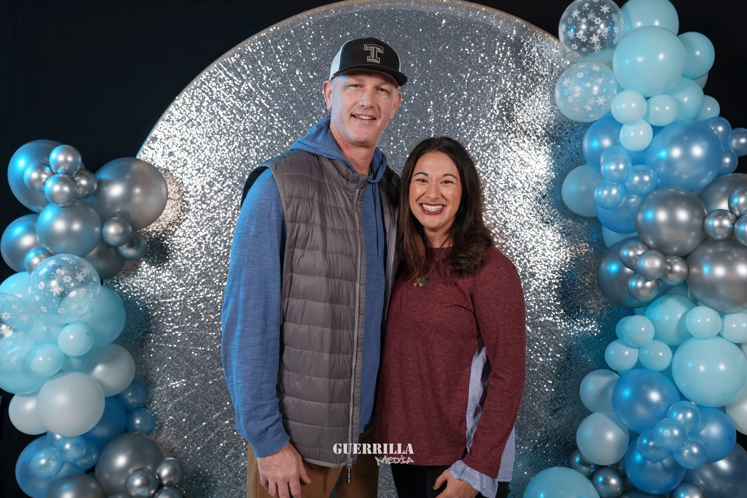 A man and woman standing side by side in front of a circular, glittery silver backdrop with blue and silver balloons on either side. They are smiling and dressed casually.