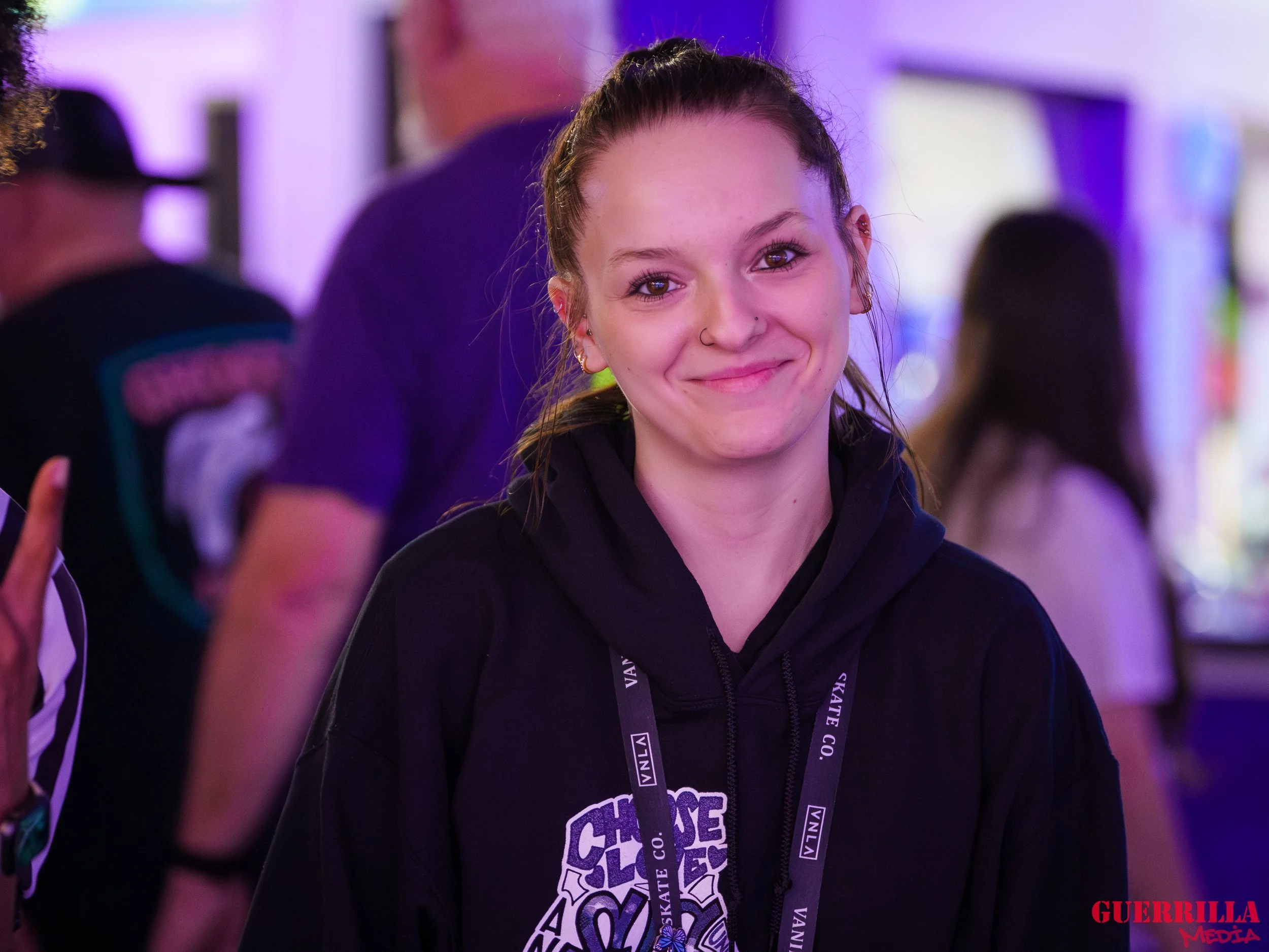 A young woman with a slight smile, wearing a black hoodie and a lanyard around her neck, at an indoor event with purple lighting, with people in the background.