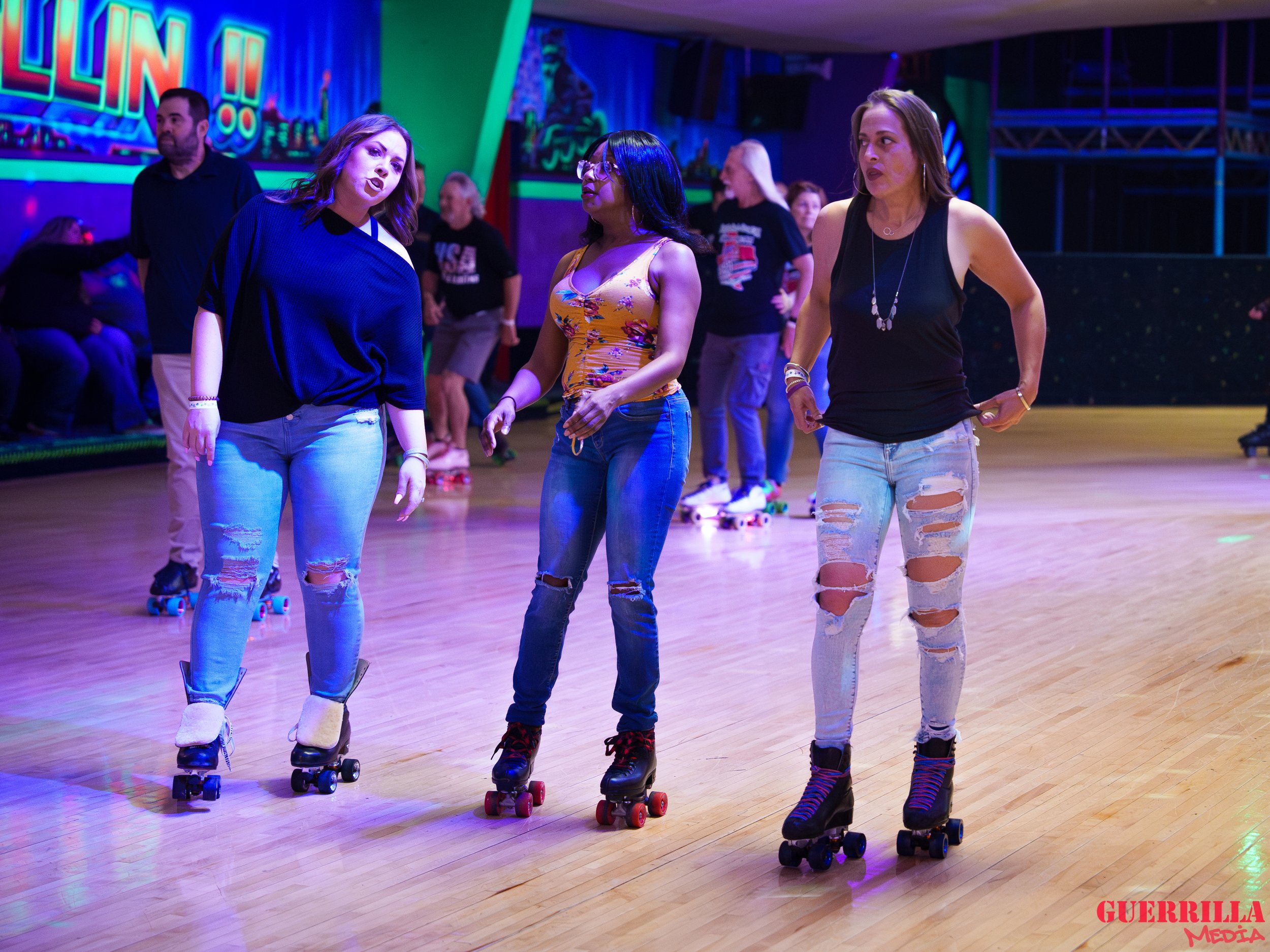 Three women roller skating together in a roller rink, engaging in conversation. They are dressed casually with ripped jeans and roller skates. Background includes other skaters and colorful neon lights.