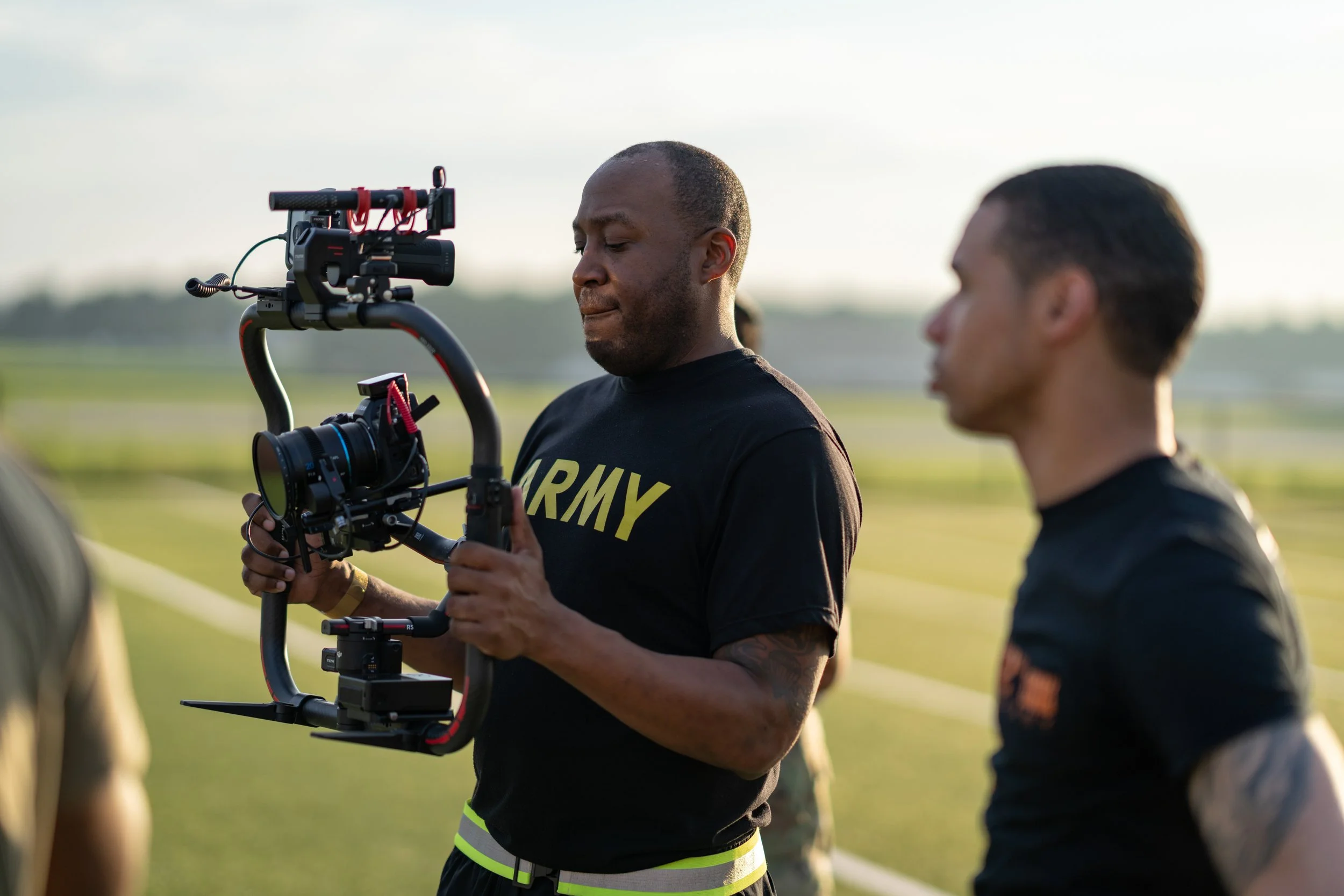 A person in army T-shirt holding a camera stabilizer with a camera attached, standing outdoors during daytime, with another person in black shirt blurred in the background.