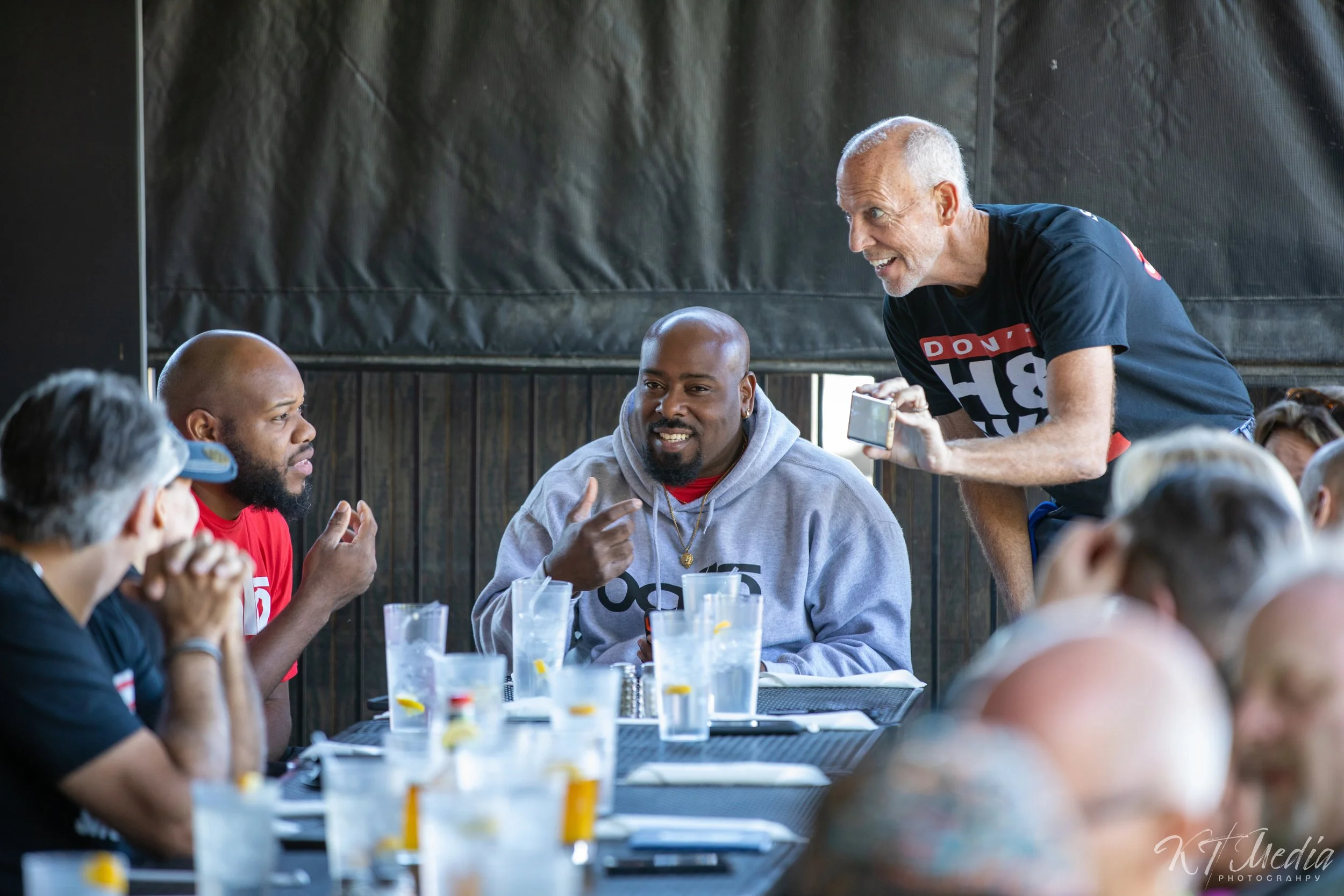 A group of people gathered around a table, engaged in conversation. One man standing up is leaning forward, holding a phone, and smiling, while the seated individuals are listening and talking. The setting appears to be a casual indoor event with gla