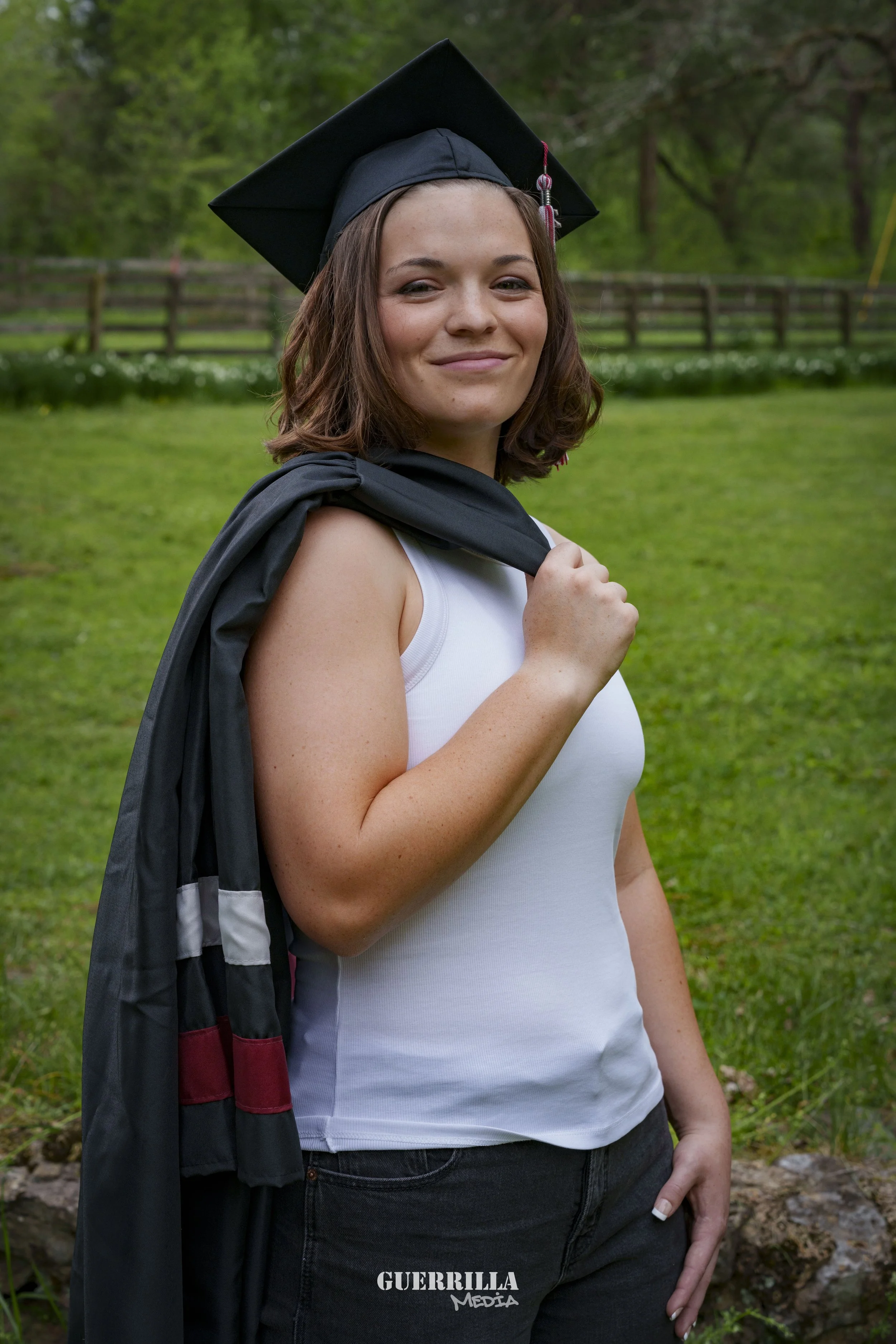 Young woman in a graduation cap and gown, holding her gown over her shoulder, standing outdoors on green grass with trees in the background.