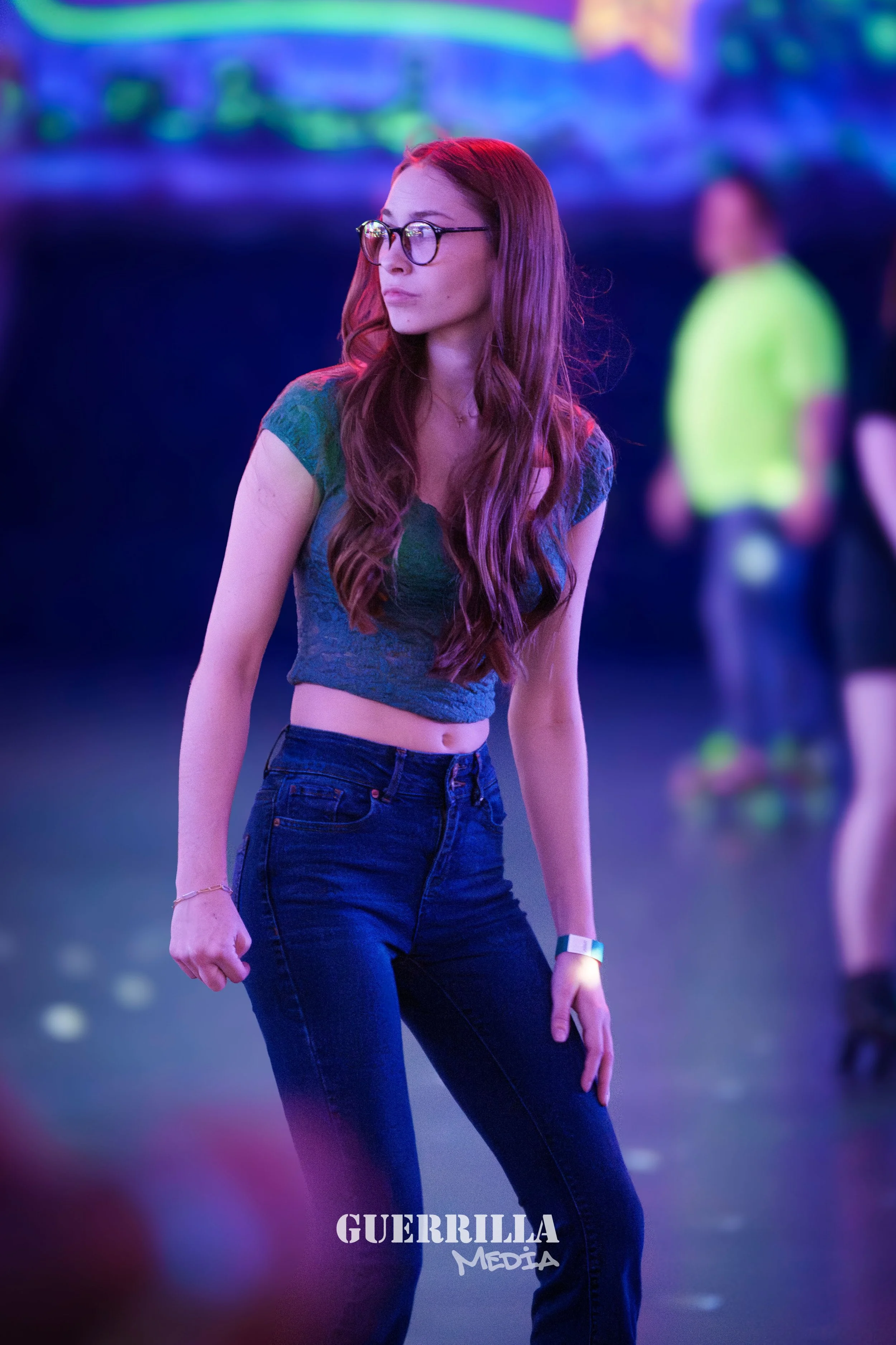 A young woman with long red hair, wearing glasses, a cropped tops, high-waisted jeans, and a fitness tracker, stands in a dimly lit space with colorful neon lights. She appears to be at a roller skating rink, with other skaters in the background.
