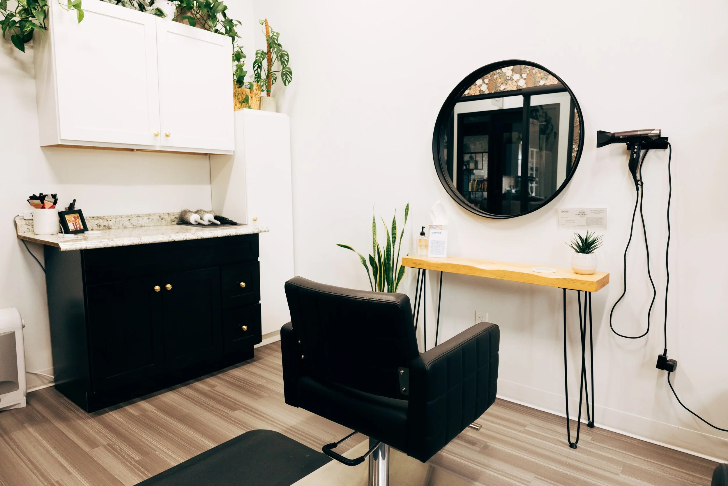 Salon with black styling chair, round mirror, wooden desk, hairdryer on wall, plants, and white cabinets.