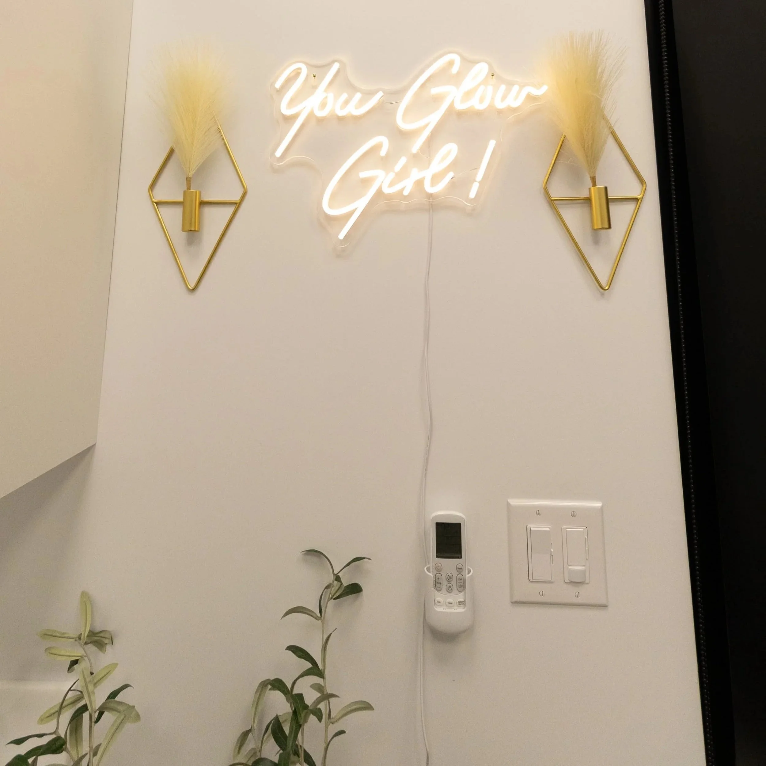 Neon sign on a white wall that reads 'You Grow Girl!' with two gold decorative wall sconces with dried pampas grass on each side of the sign.