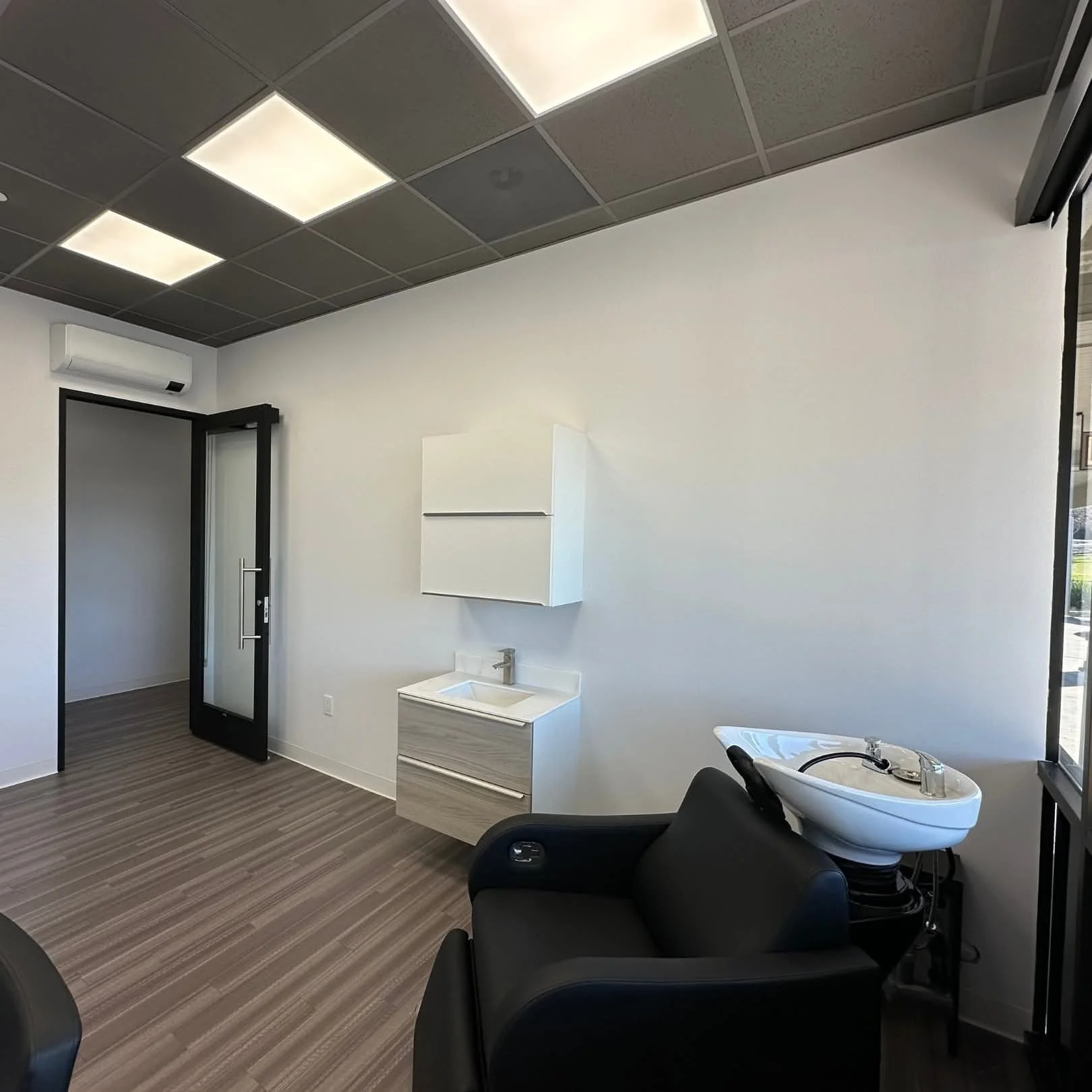 Interior of a salon or beauty studio with a hair wash station, black armchair, white wall-mounted cabinet, small sink with a faucet, wood laminate flooring, and ceiling lights.
