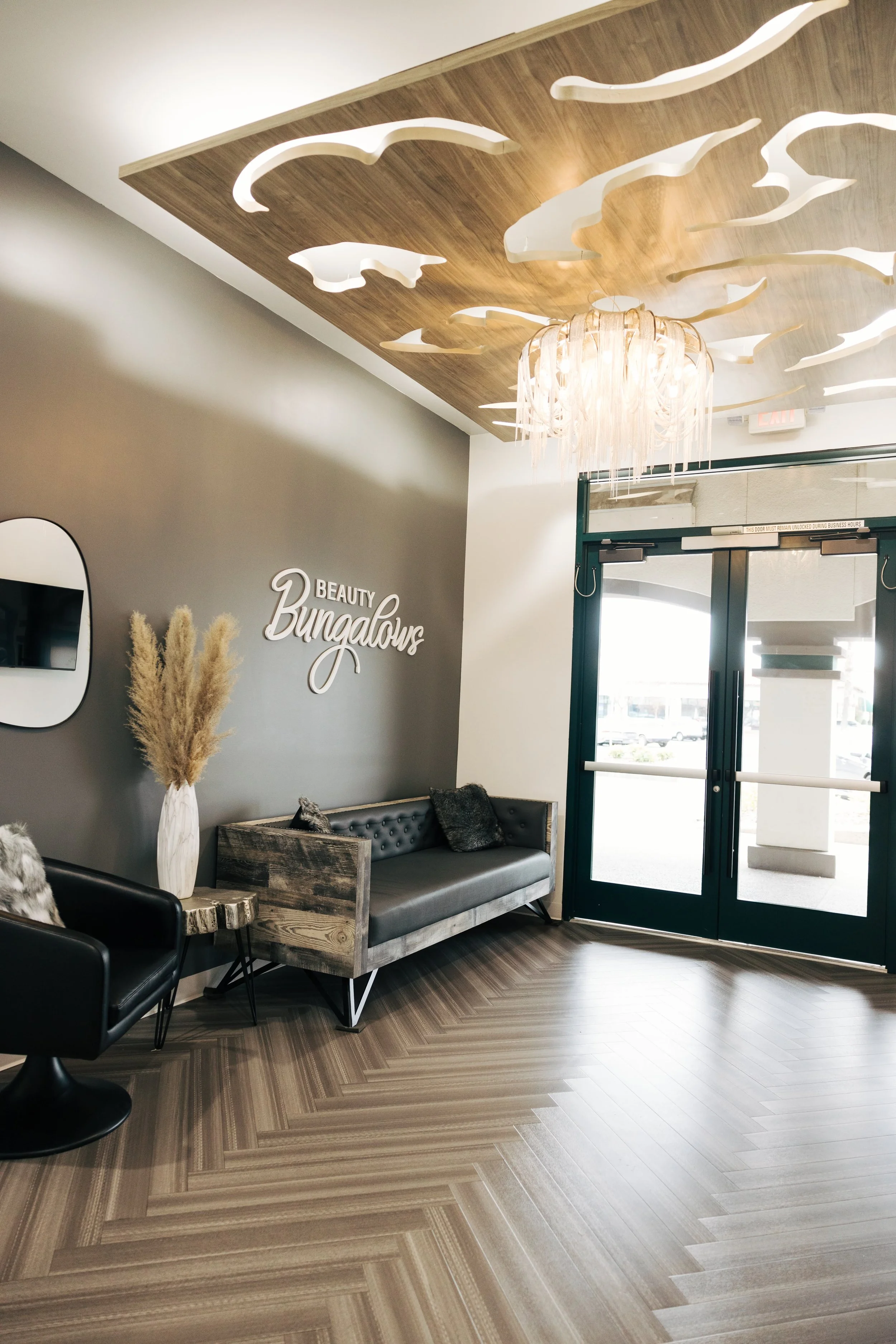 Interior lobby area of a beauty salon called 'Beauty Bungalou' with modern decor, a black leather chair and a wooden bench with cushions, a vase with pampas grass, and large glass doors at the entrance.
