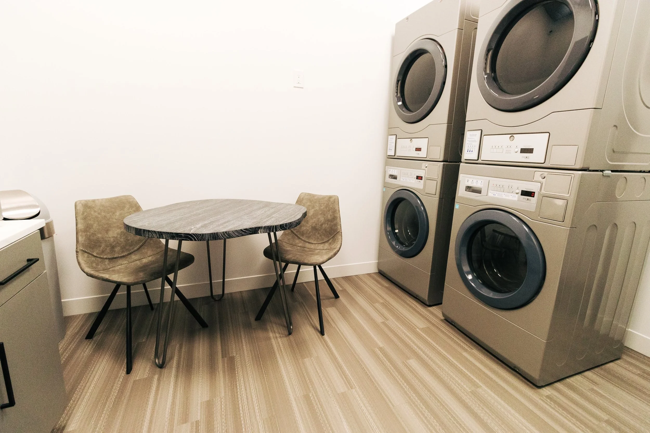 A laundry room with four stacked washer and dryer units on the right, a small round table with two tan chairs on the left, and a countertop with a radio on it. The room has bamboo flooring and white walls.