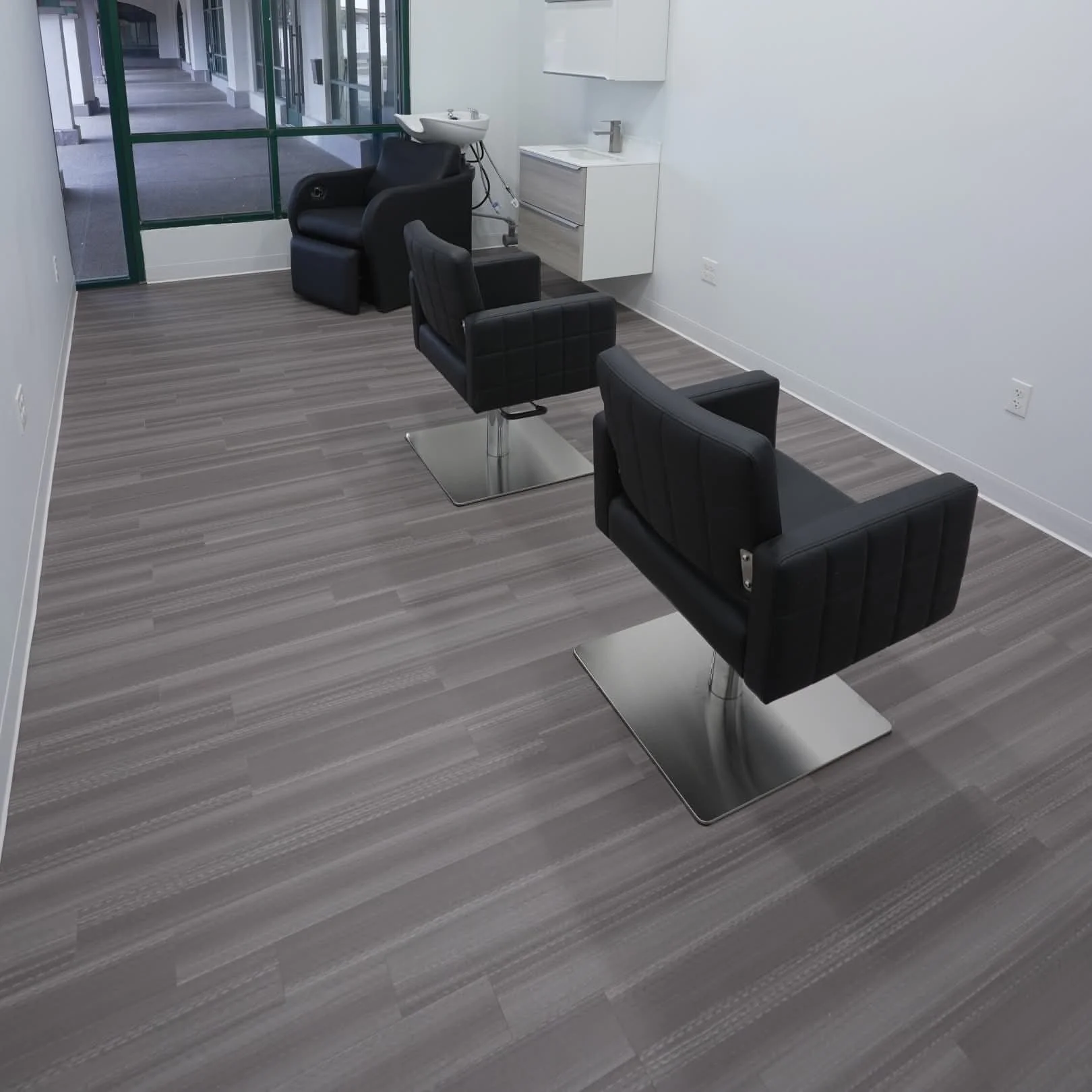 Empty hair salon with three black styling chairs, a wash basin, and a cabinet, with large windows showing an outdoor corridor.