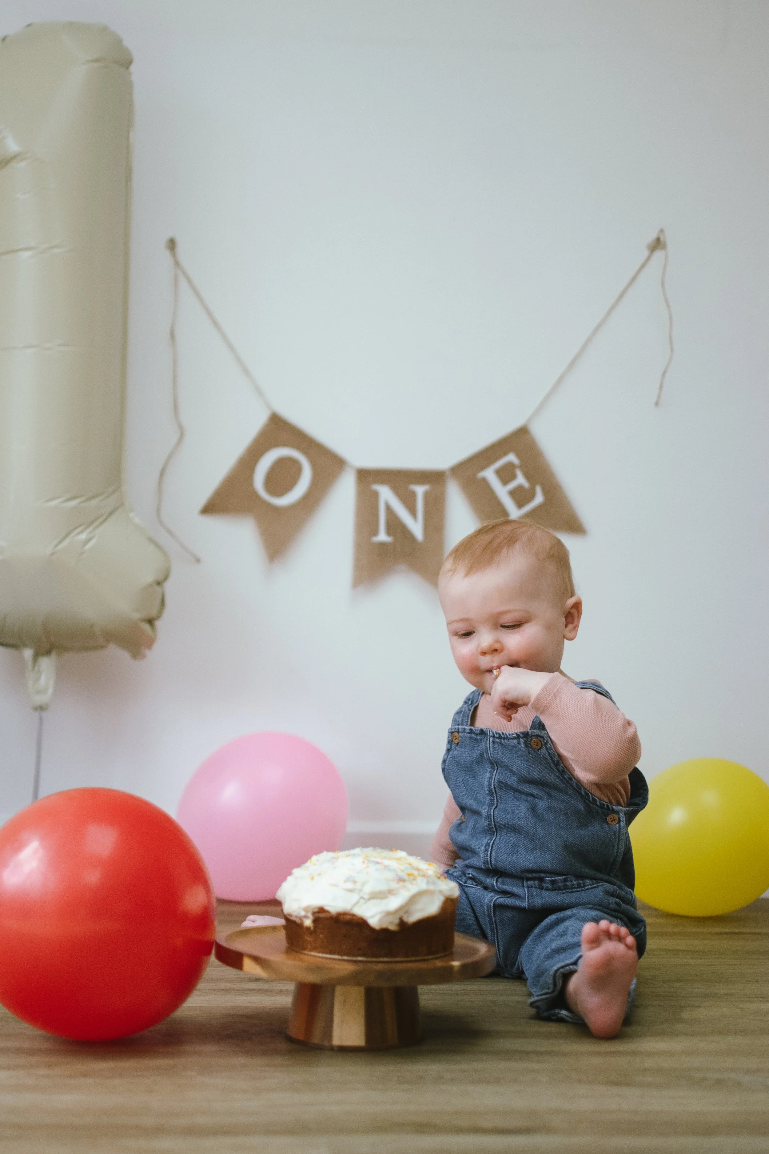 A young boy sitting on a wooden floor at a birthday celebration, surrounded by colorful balloons, with a birthday cake in front of him, a 'ONE' banner on the wall behind him, and a balloon shaped like the number 1 on his left.