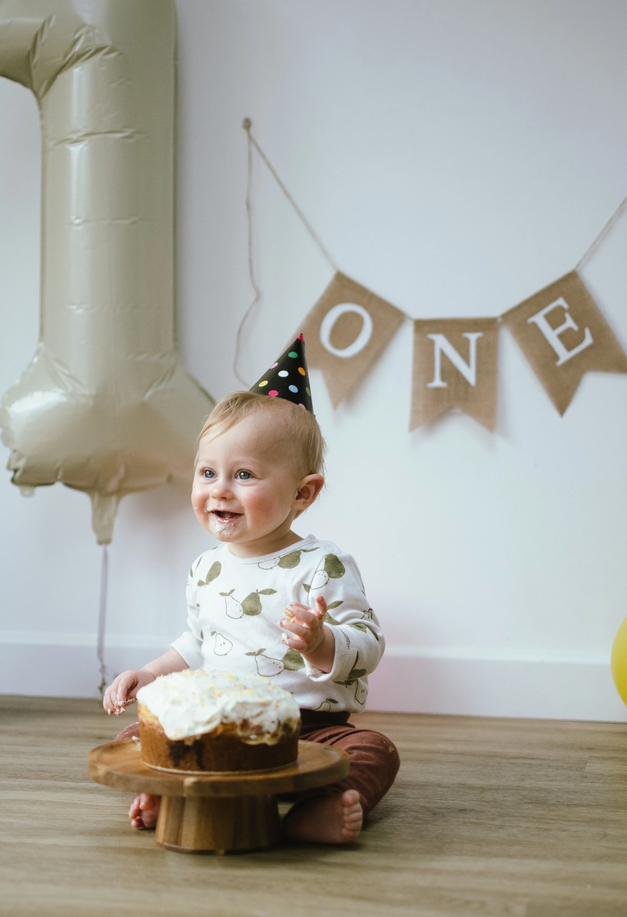 Smiling young child celebrating birthday, wearing a party hat, sitting on the floor with a loaf cake with icing and sprinkles, birthday decorations in background including a large balloon and a 'ONE' banner.