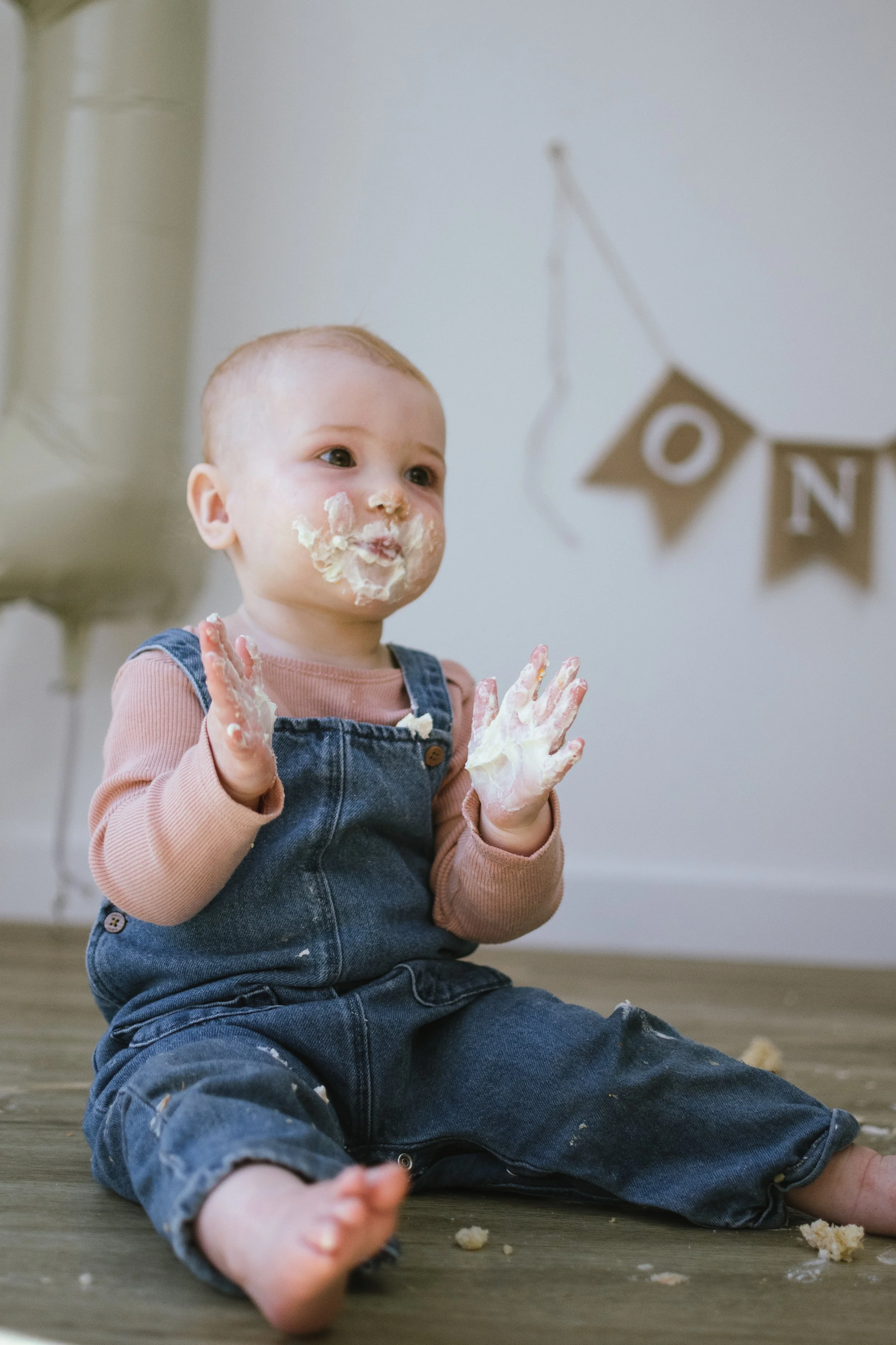A young child sitting on a wooden floor with food and cake in their hands, face, and clothes, with a cake and bunting in the background.