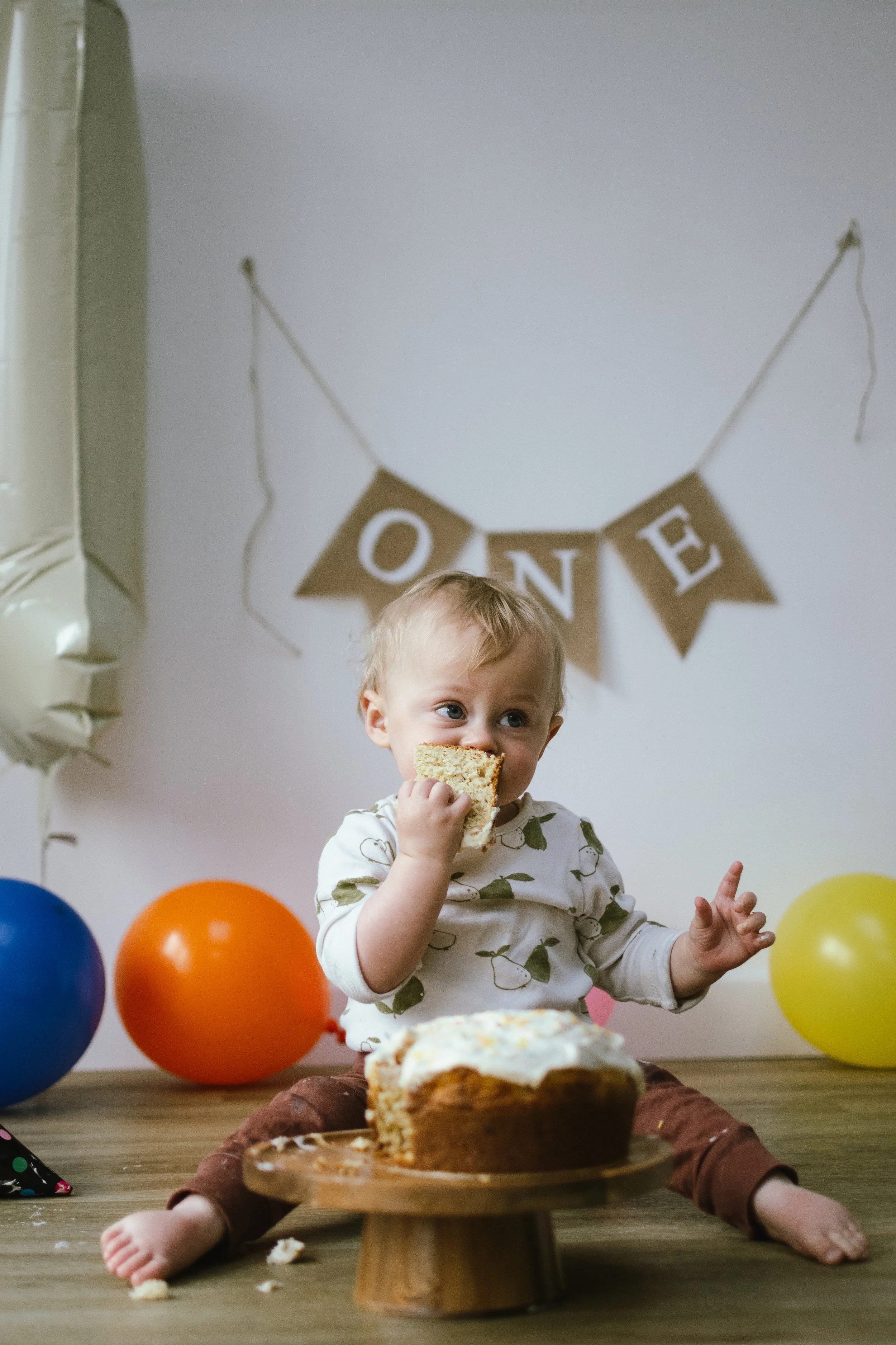 A young child sitting on a wooden floor at a birthday party, eating cake with colorful balloons and a "ONE" banner in the background.