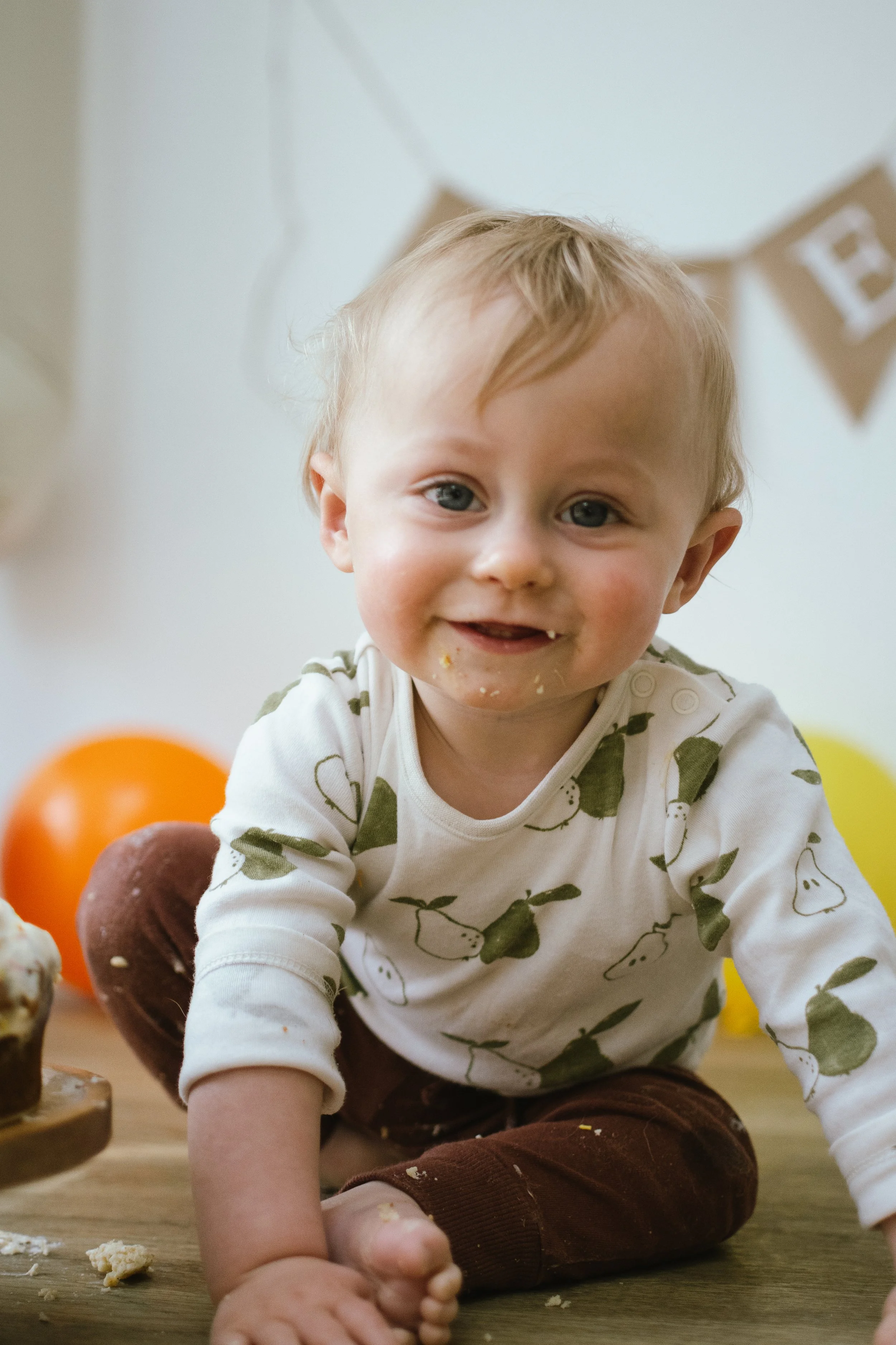 A young child with blond hair, blue eyes, and a toothy smile, sitting on the floor, surrounded by colorful balloons and cake, wearing a white shirt with green pear patterns and brown pants.