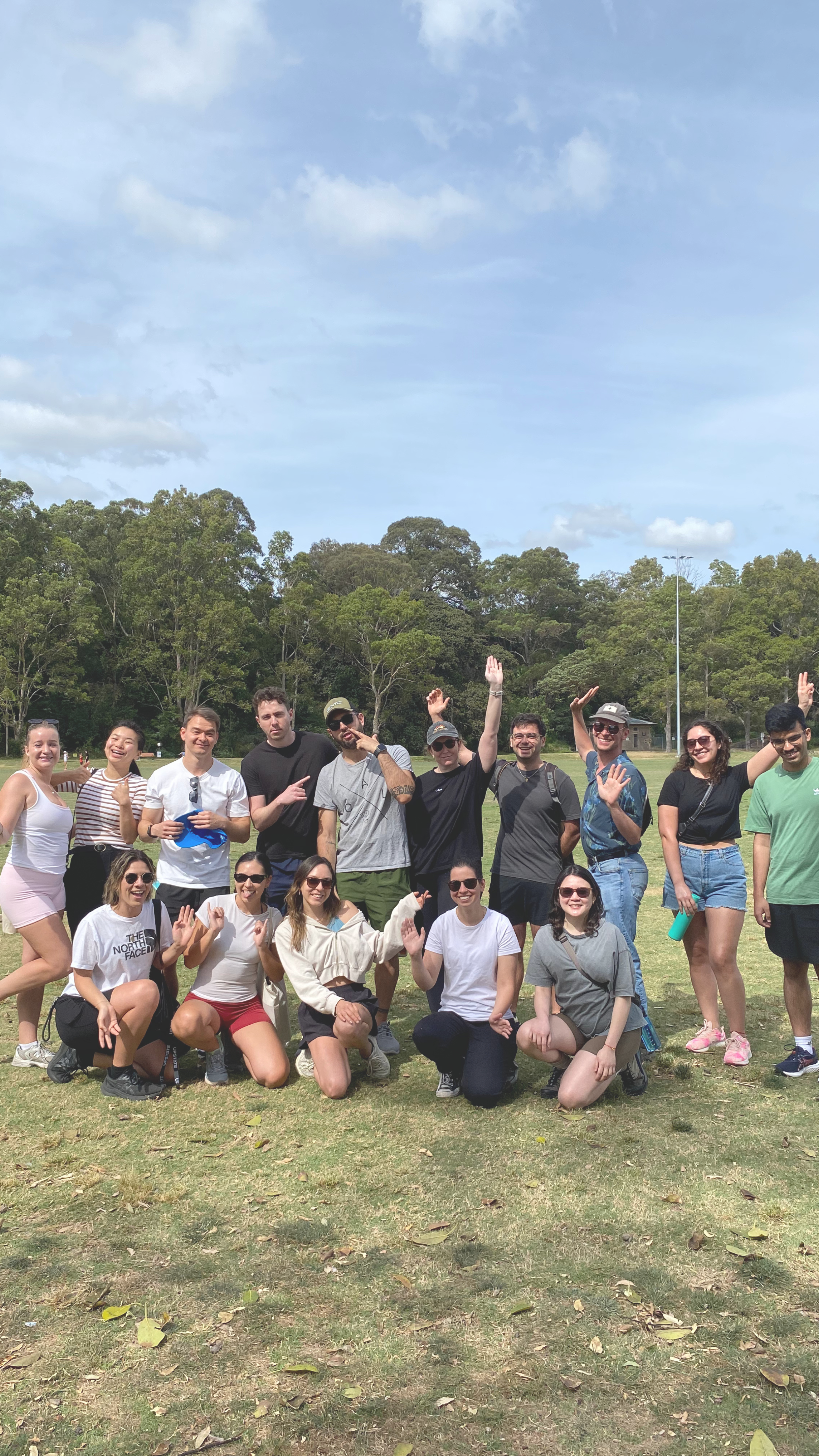 Group of people posing outdoors on a grassy field with trees in the background, some standing and some kneeling, smiling and waving.
