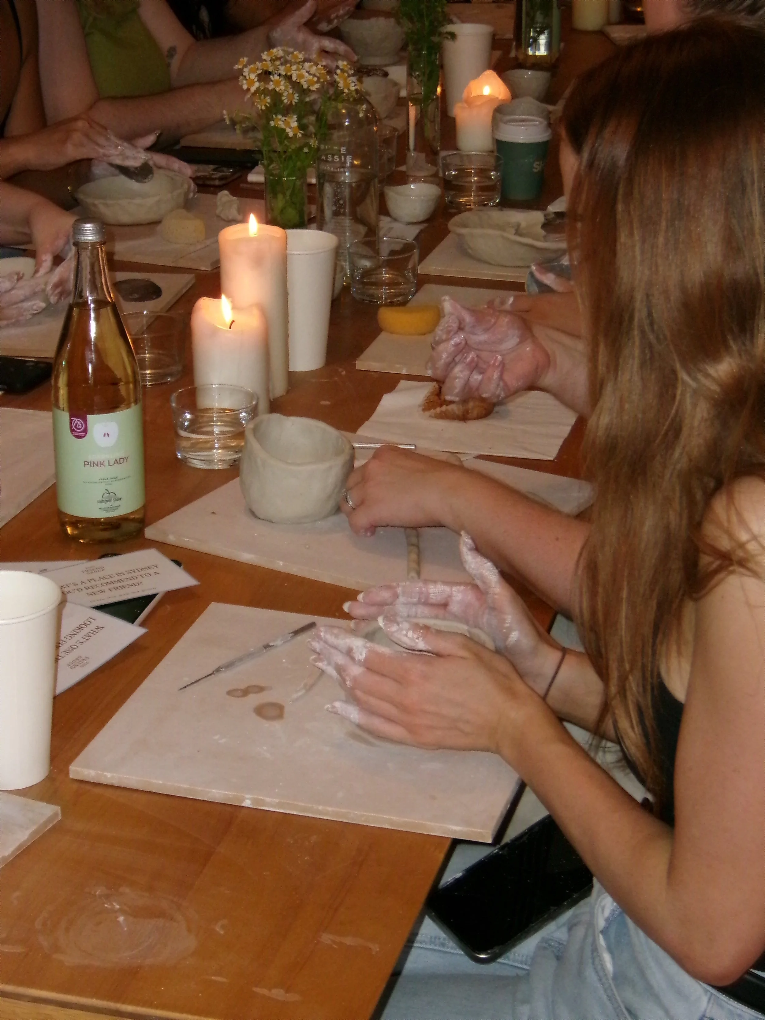 People participating in a pottery class, shaping clay on pottery wheels at a wooden table with lit candles and various drinks.