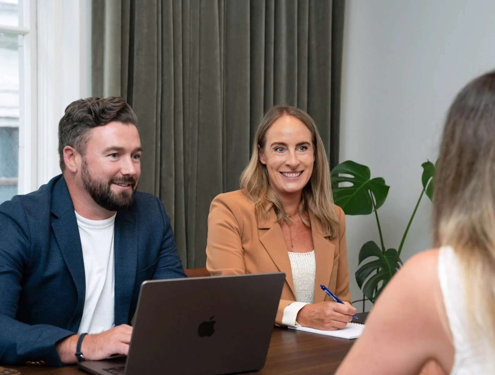 Two professionals, a man with a beard and a woman with blonde hair, sit at a table in a meeting room talking with a woman across from them. The woman is smiling, holding a pen, and has a notebook in front of her. There is a laptop in front of the man, and a large green plant in the background.