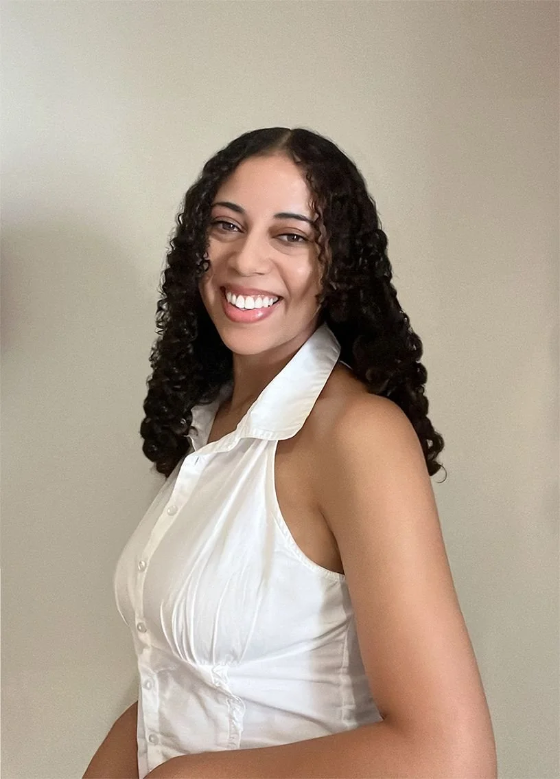 A woman with curly dark hair, smiling, wearing a sleeveless white blouse, standing against a beige background.