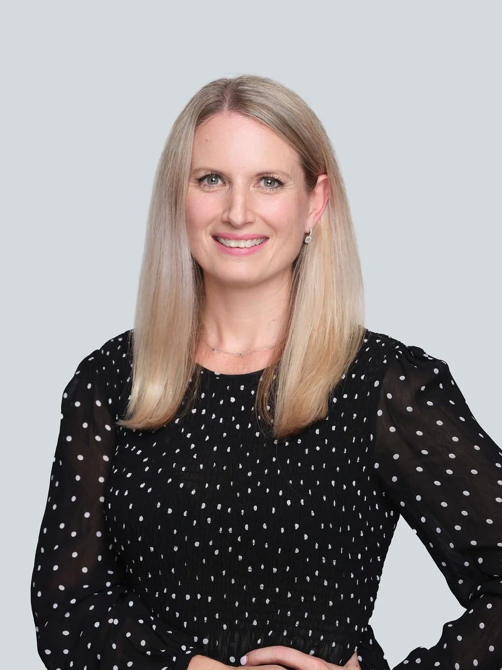 Professional woman with blonde hair wearing a black dress with white polka dots, smiling against a light background.