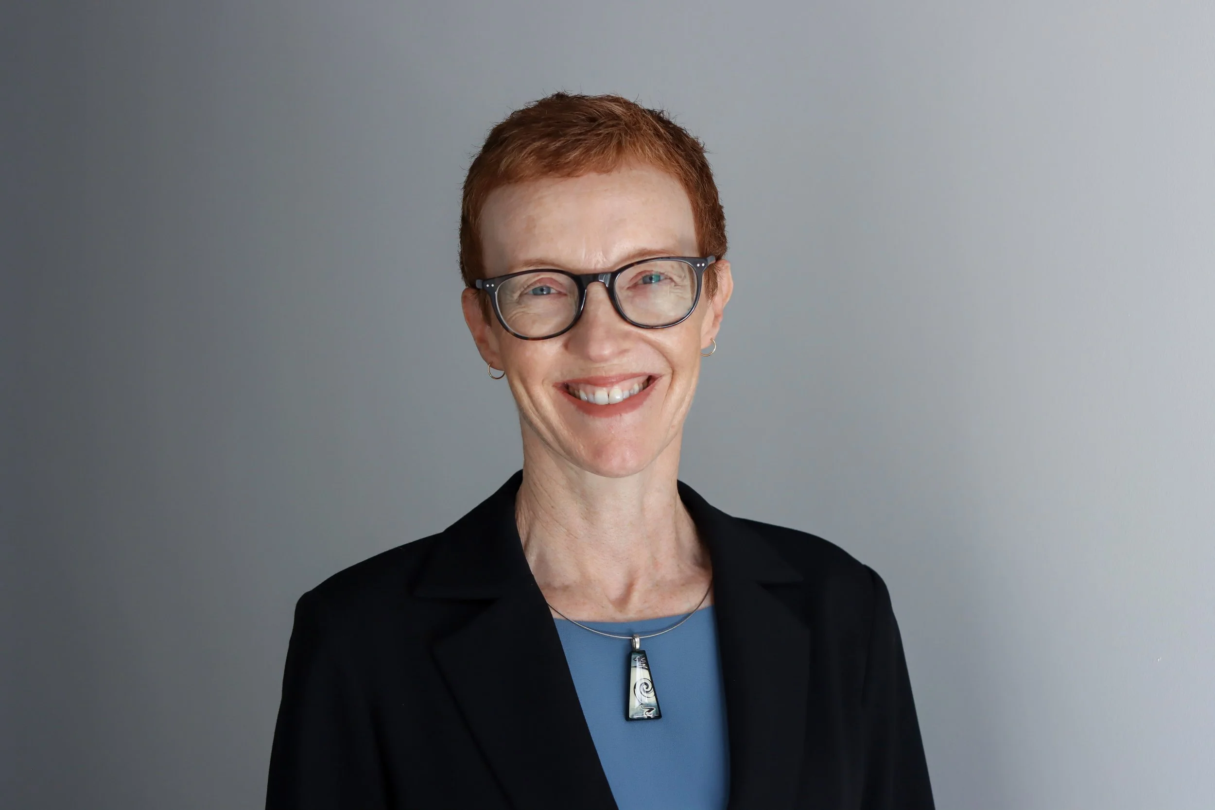 A smiling woman with short red hair, glasses, and earrings, wearing a black blazer and blue top, standing against a plain gray background.