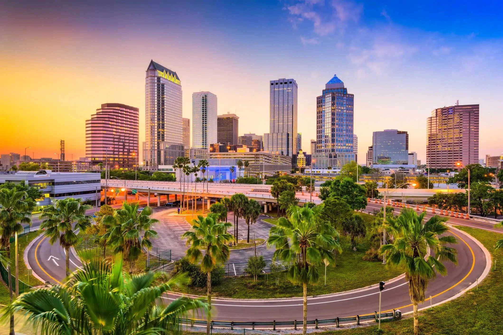 Colorful sunset over Tampa’s city skyline with palm trees and highways in the foreground.
