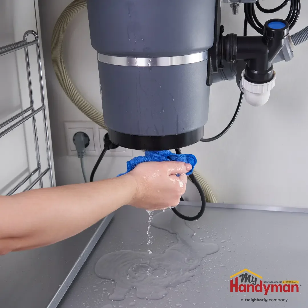 A hand using a blue microfiber cloth to wipe the underside of a gray garbage disposal unit beneath a kitchen sink, with water dripping, in a Saline, Michigan home.