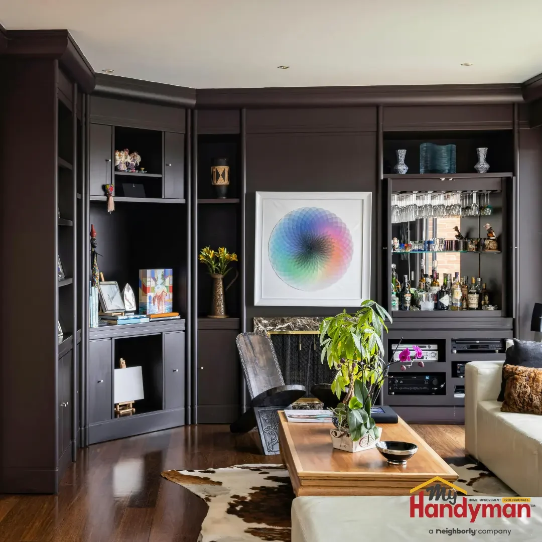 A living room with dark floor-to-ceiling custom built-in shelving and cabinetry displaying books, glassware, and decor around a framed artwork in an Ann Arbor, Michigan home.