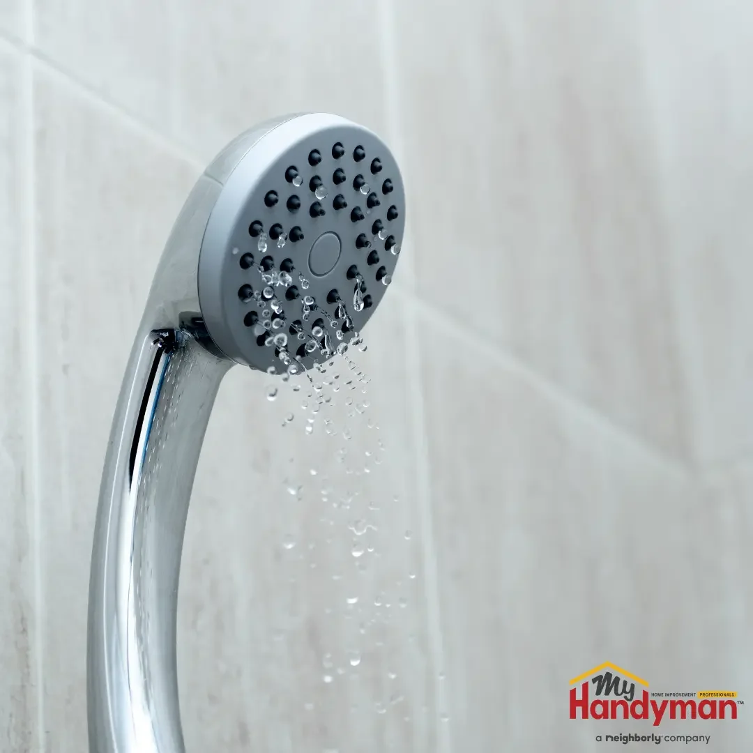A chrome handheld showerhead with a gray nozzle face running a thin, reduced spray against a tiled wall in a Saline, Michigan bathroom.