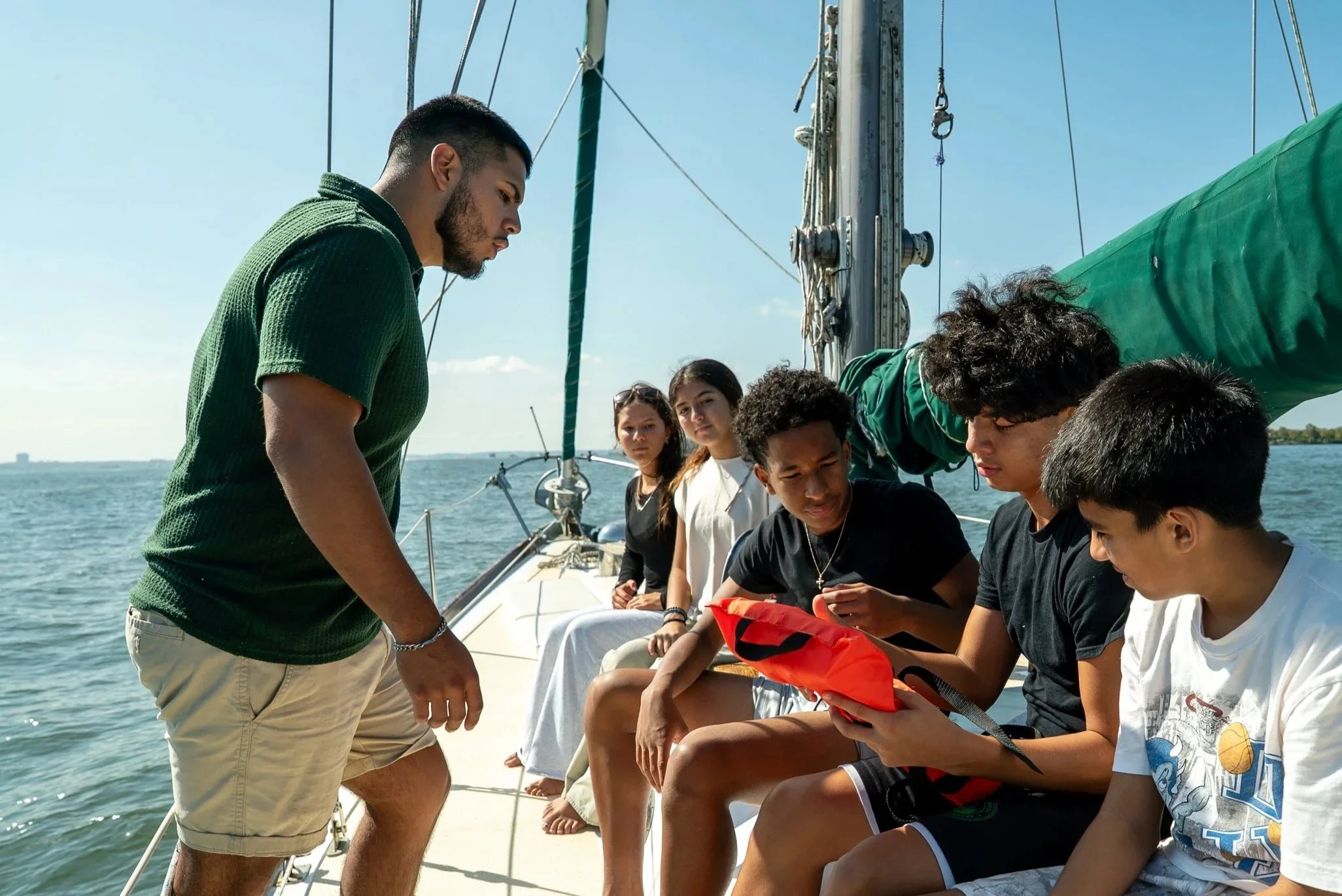 Young man instructing a group of teenagers on a sailboat, with water and a clear sky in the background.