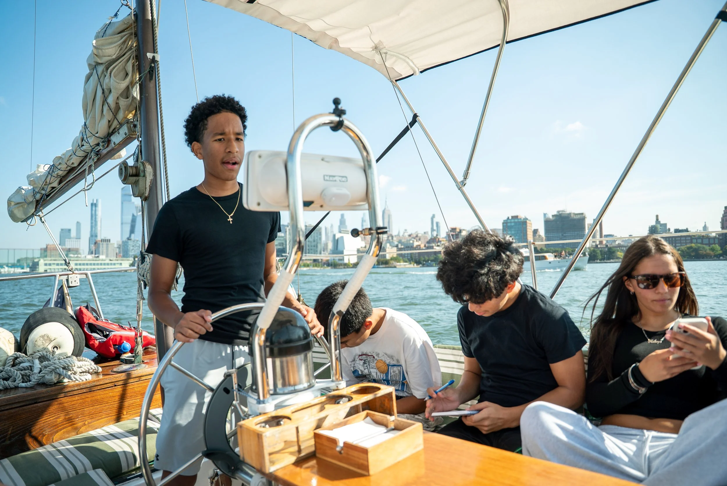 A group of young people on a boat, with a city skyline in the background, some are looking at their phones and a young man is steering the boat.
