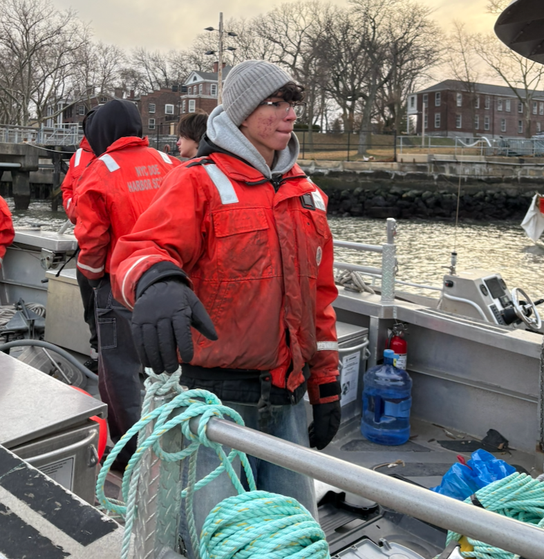 A young person wearing a gray beanie, glasses, a red jacket with reflective strips, and gloves stands on a boat, holding a coiled rope with water and houses in the background.