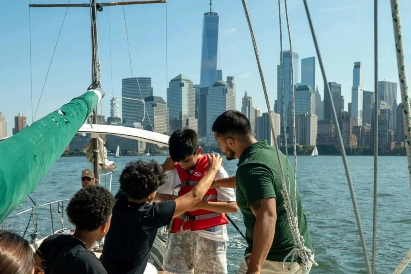 A group of people on a sailboat in New York City, with the skyline including One World Trade Center in the background. One person, a young boy, is receiving assistance from adults while wearing a life jacket.