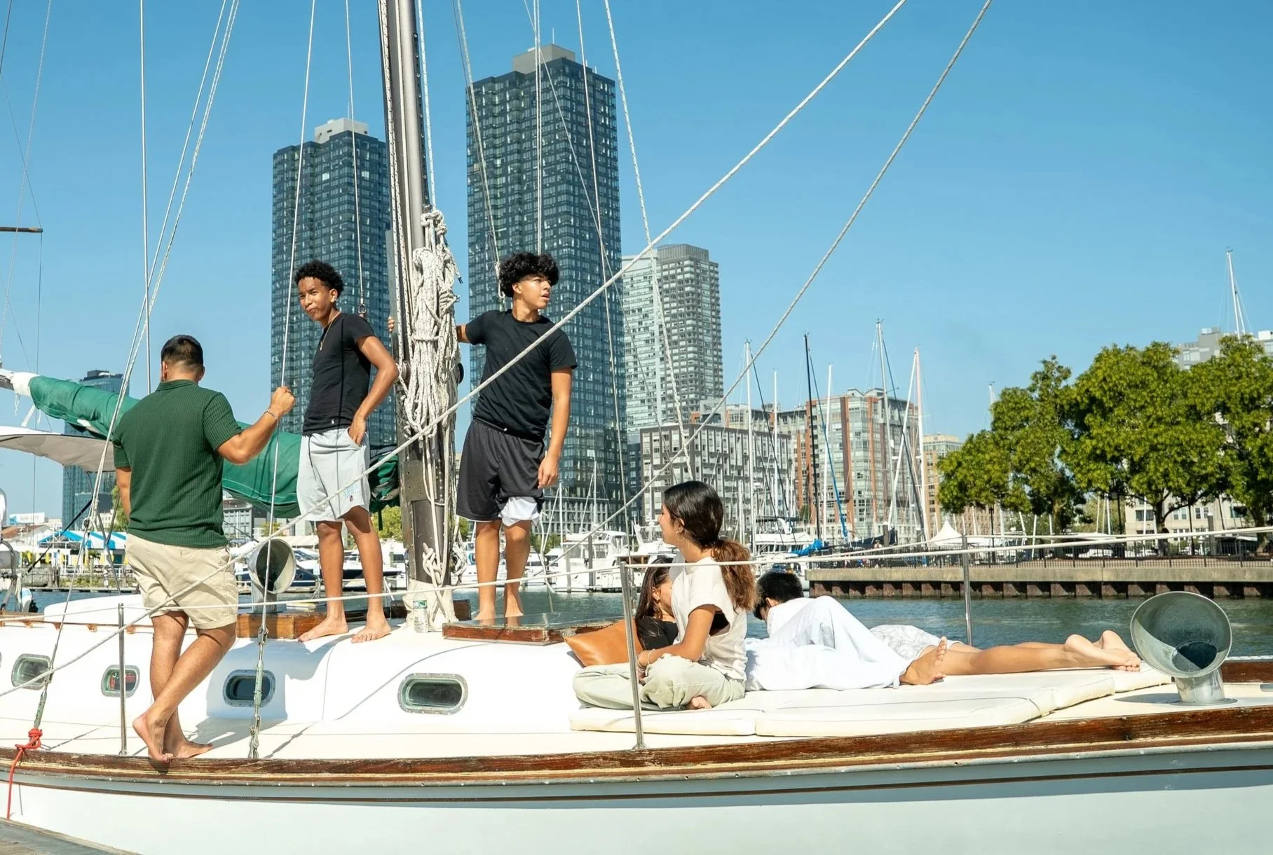 A group of young people on a sailboat docked at a marina, with tall city buildings and trees in the background. Some are sitting, some are standing, engaged in conversations and preparing for sailing.