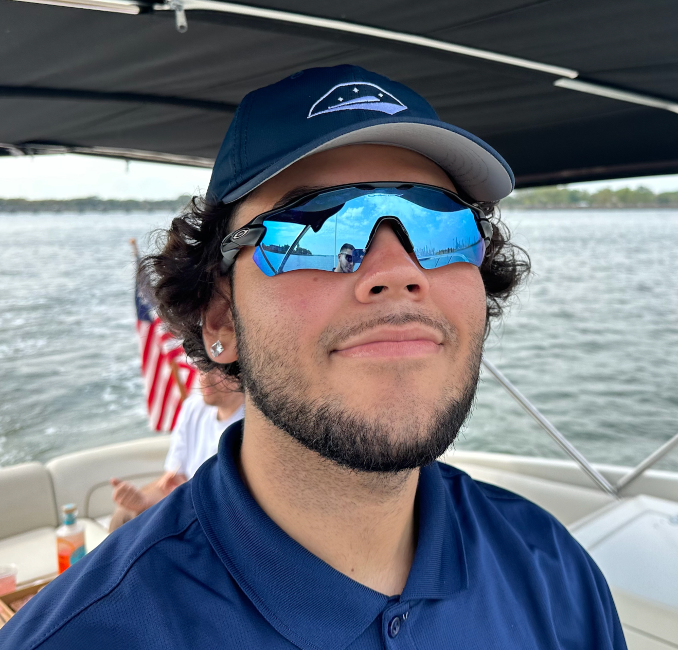 A young man with dark curly hair, wearing a navy blue polo shirt, a blue cap with a white brim and logo, and reflective blue sunglasses, smiling on a boat with water and an American flag in the background.
