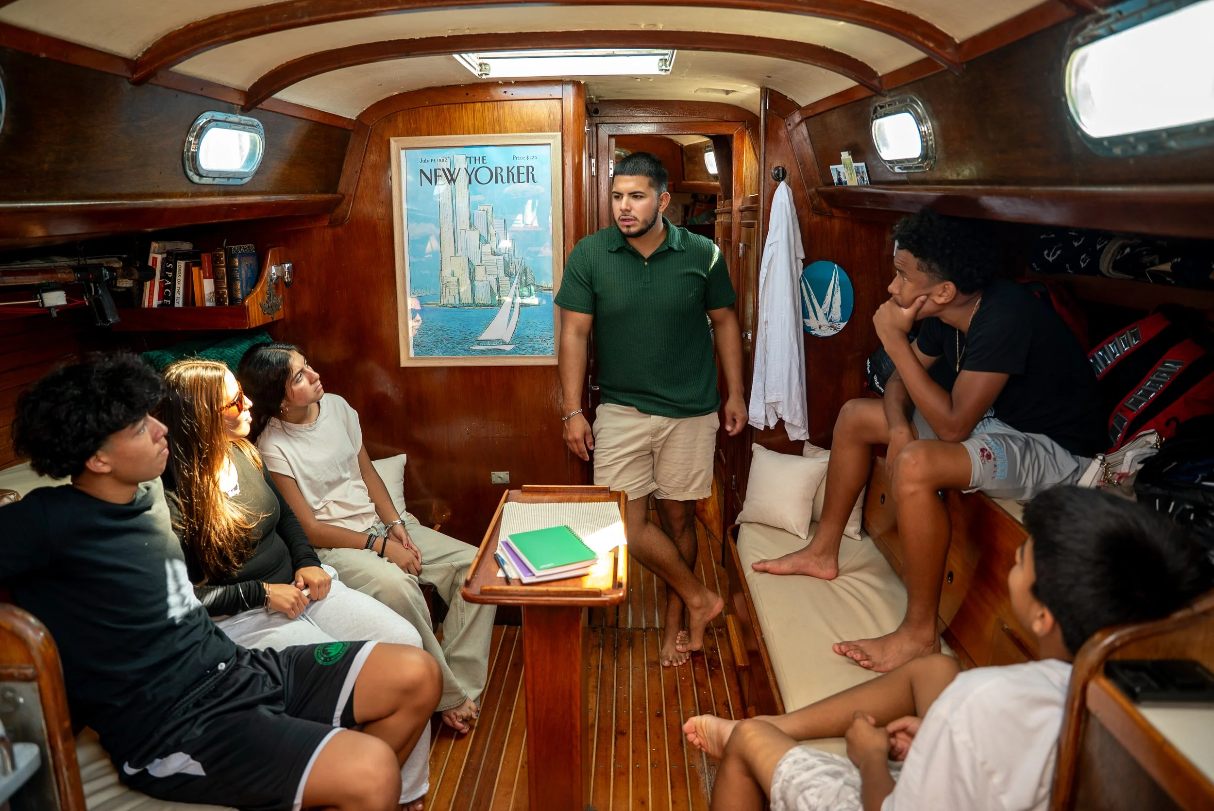 People sitting inside a wooden boat with a man standing and speaking to a group of young adults.