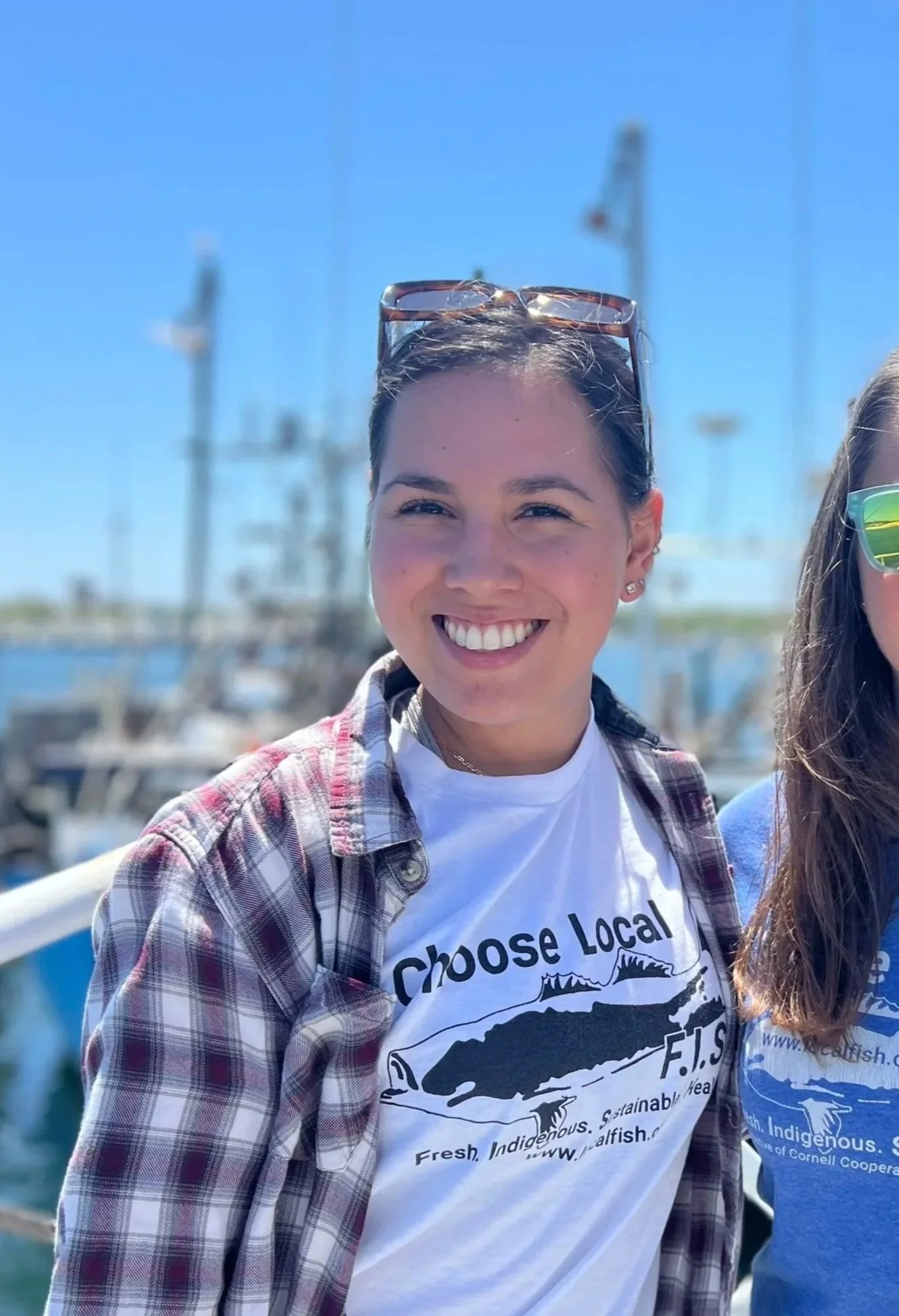 Smiling woman wearing sunglasses on her head and a plaid shirt over a white California fish advocacy T-shirt, standing outdoors near water with sailboats and masts in the background.