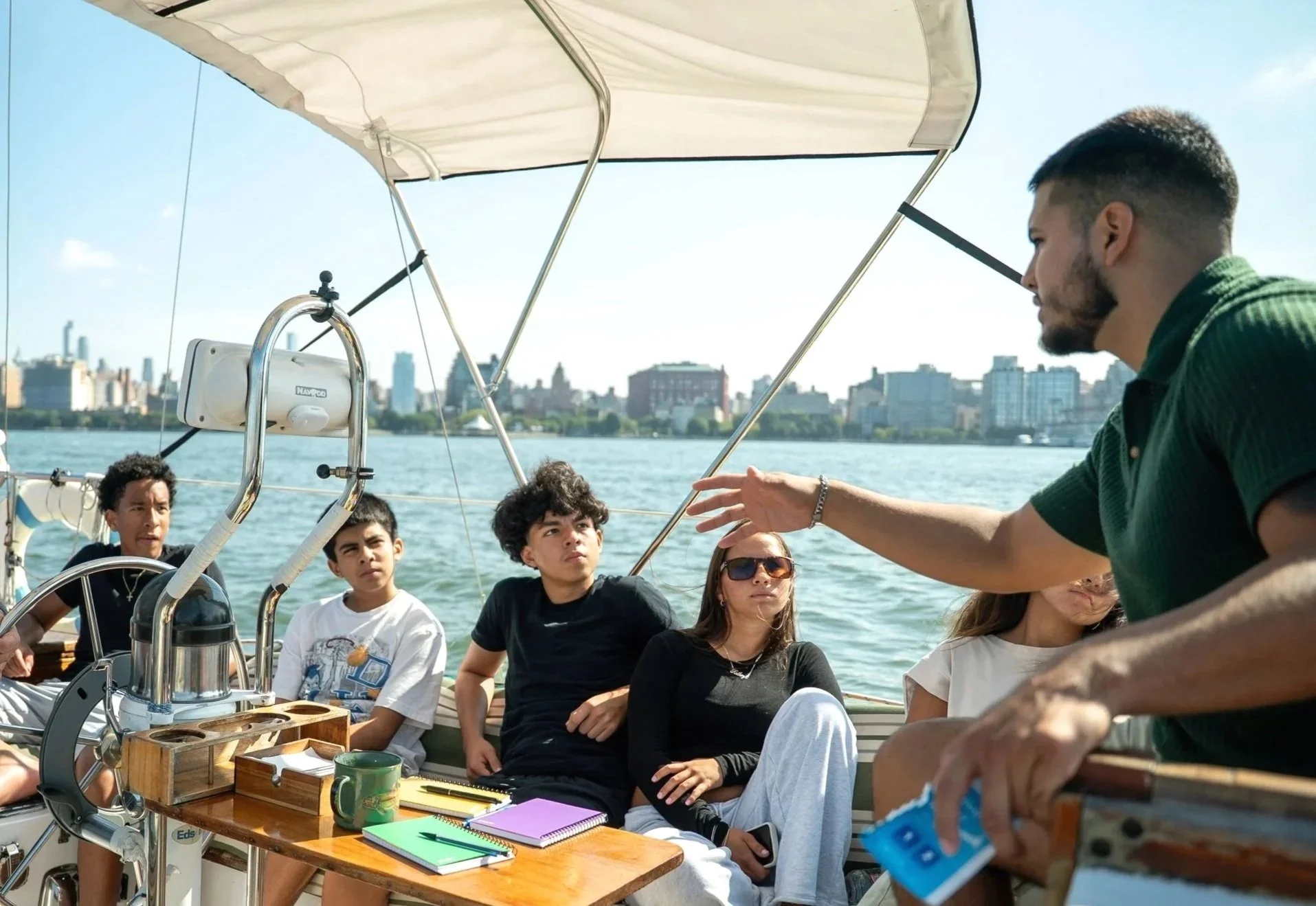 A group of young people sitting on a boat, listening to a man who is speaking and gesturing with his hand, with a city skyline in the background.