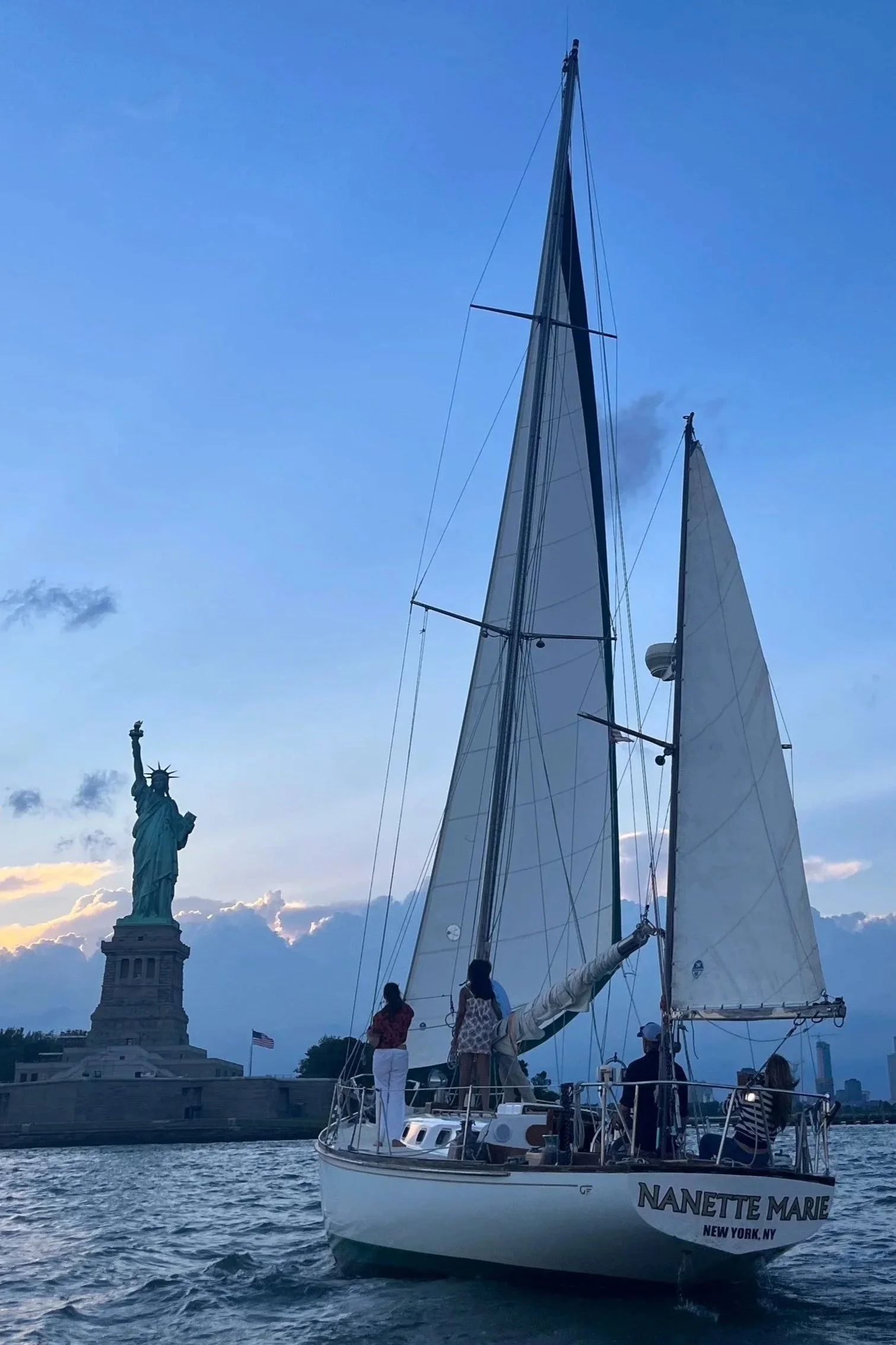 A sailboat named Nanette Marie sailing near the Statue of Liberty in New York Harbor during sunset, with several people on board and the New York City skyline in the background.