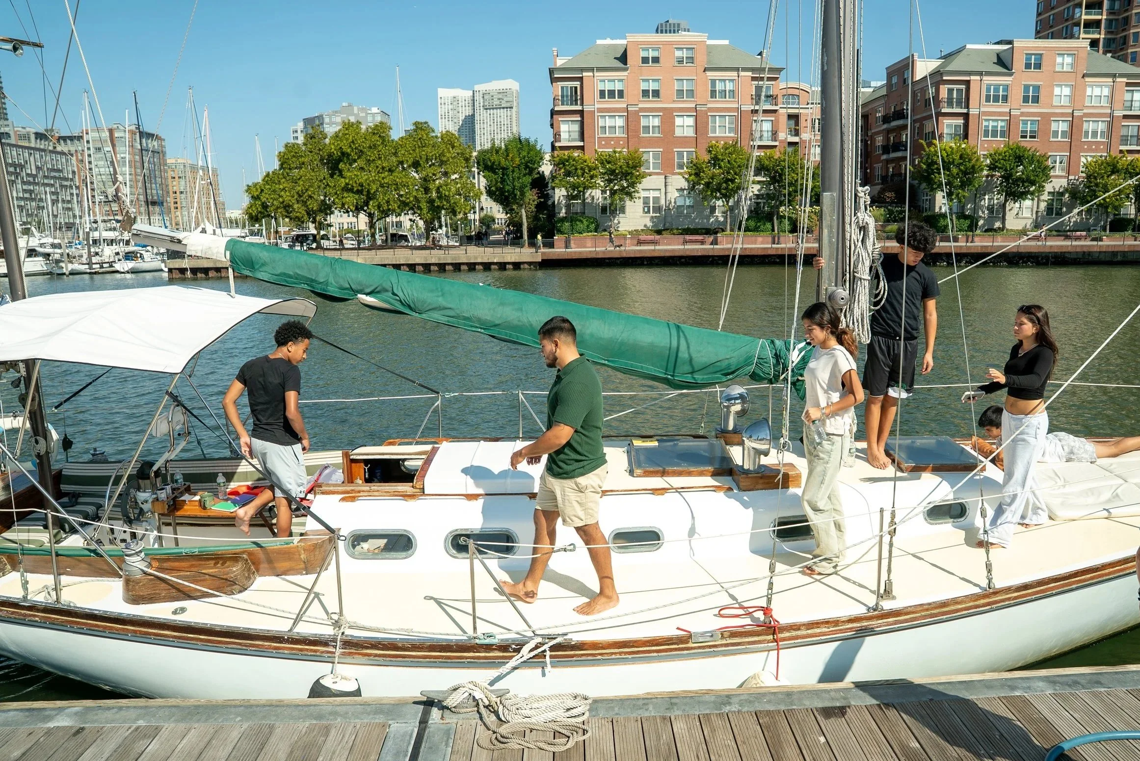 Group of five people preparing a sailboat for sailing on a marina with water and city buildings in the background on a clear day.