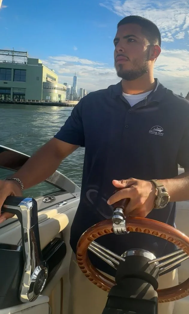 Man with a beard steering a boat on a river with city skyline in the background, wearing a navy polo shirt and a watch.