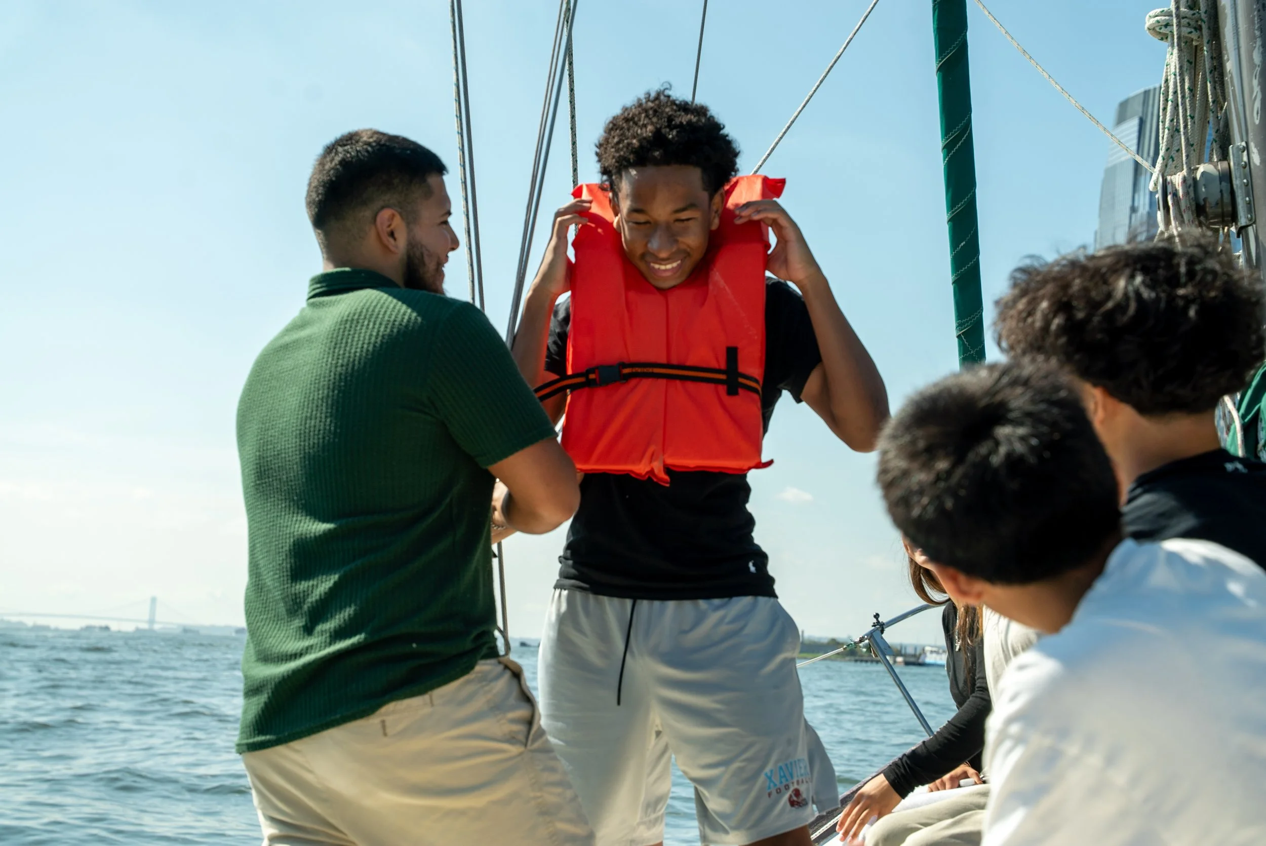 A group of young people on a boat, smiling, with one person putting on a life jacket among friends during daytime sailboat outing.