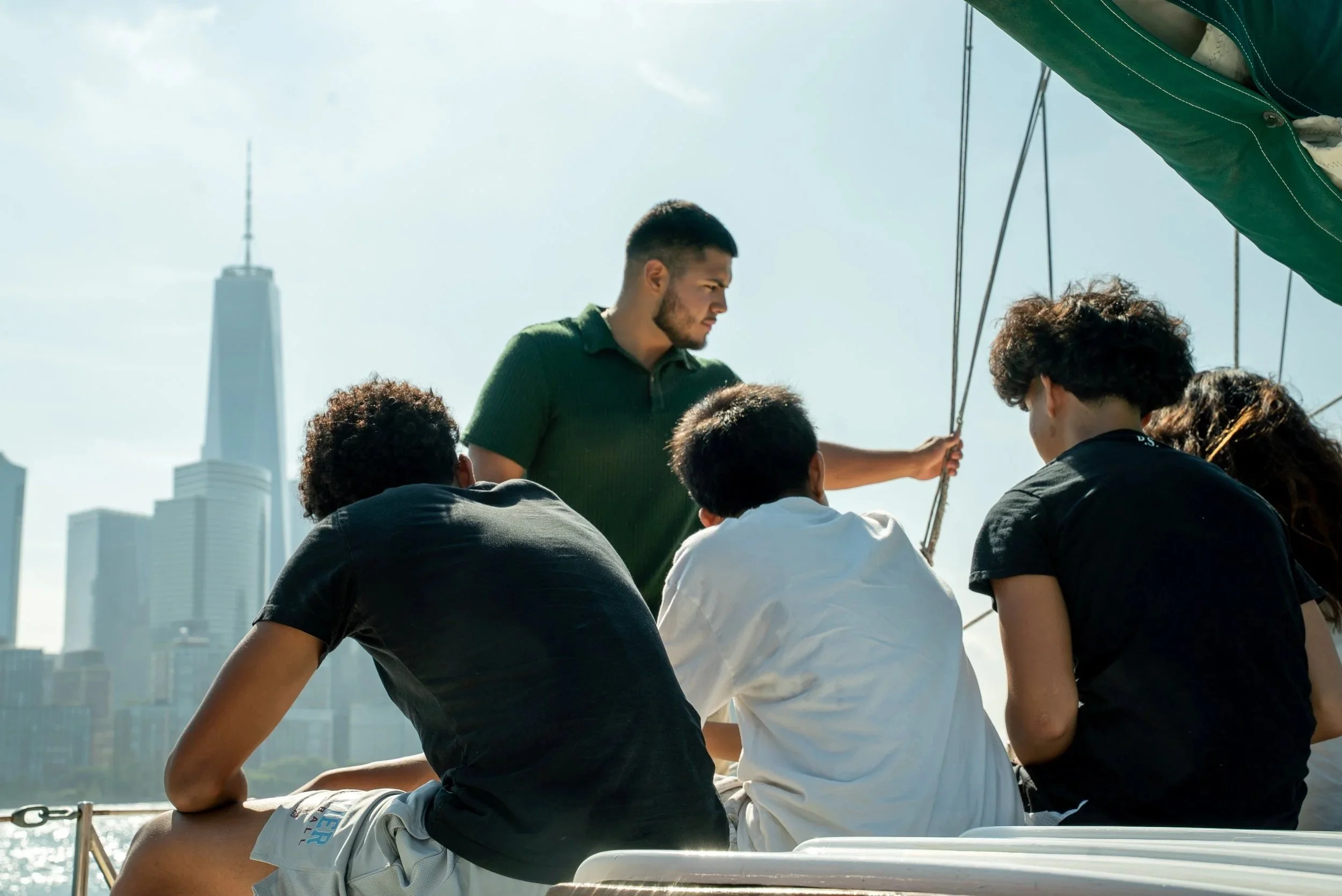 A group of young people on a boat, sitting together and talking, with a city skyline featuring tall buildings and a tower in the background.