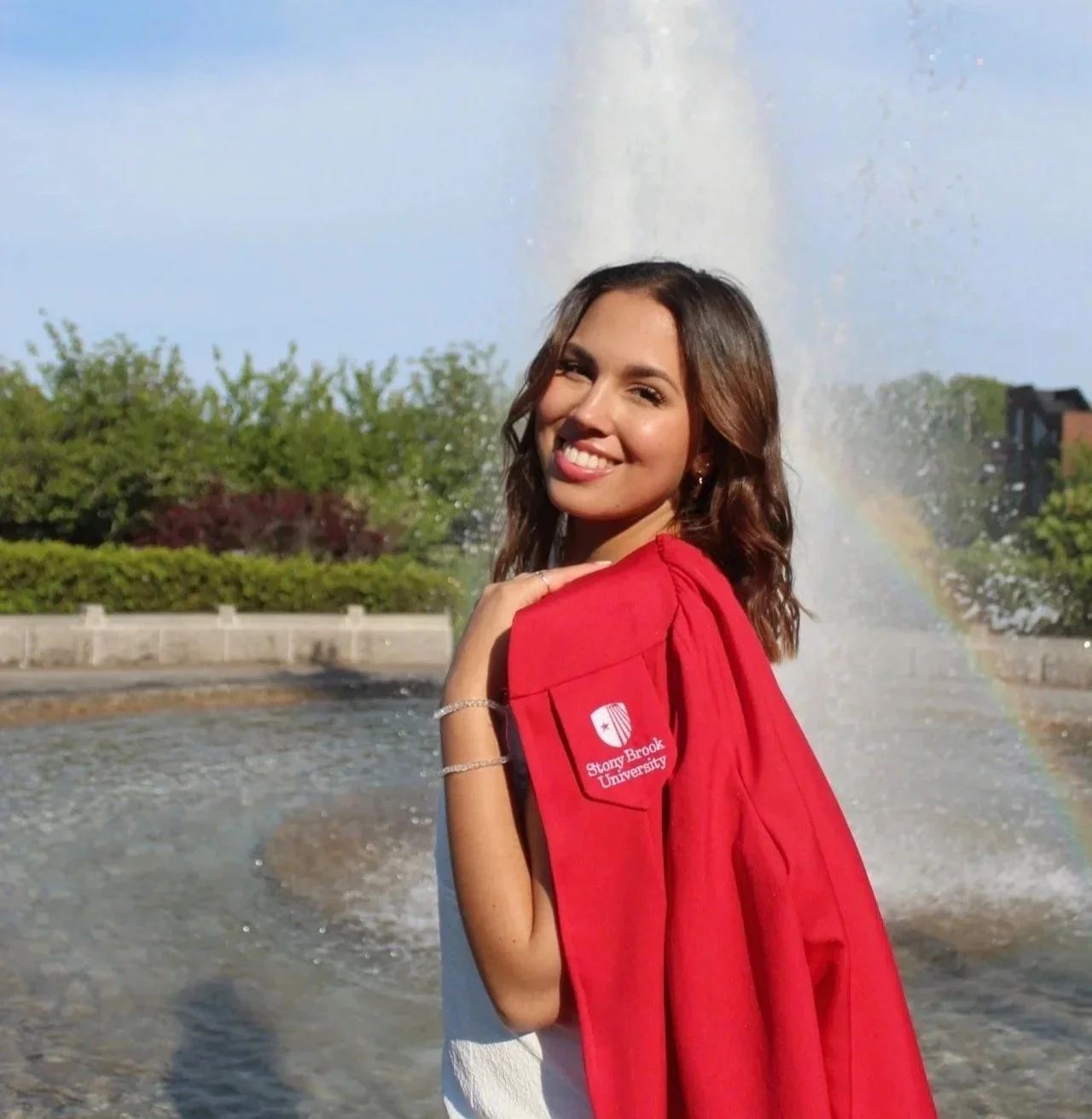 A young woman in a red graduation gown standing near a fountain with a rainbow in the background, smiling at the camera.
