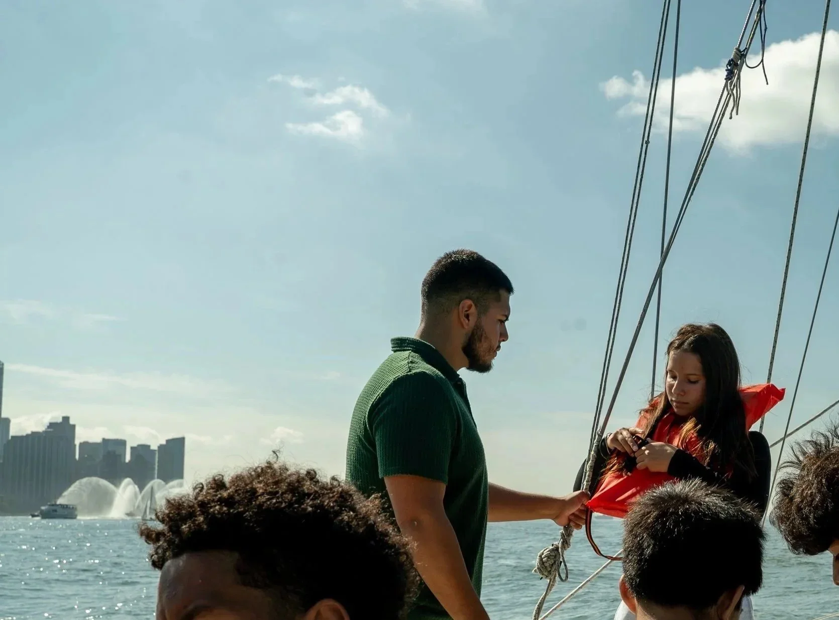 A man helps a young girl fasten her life jacket on a boat with a city skyline and water fountain in the background.