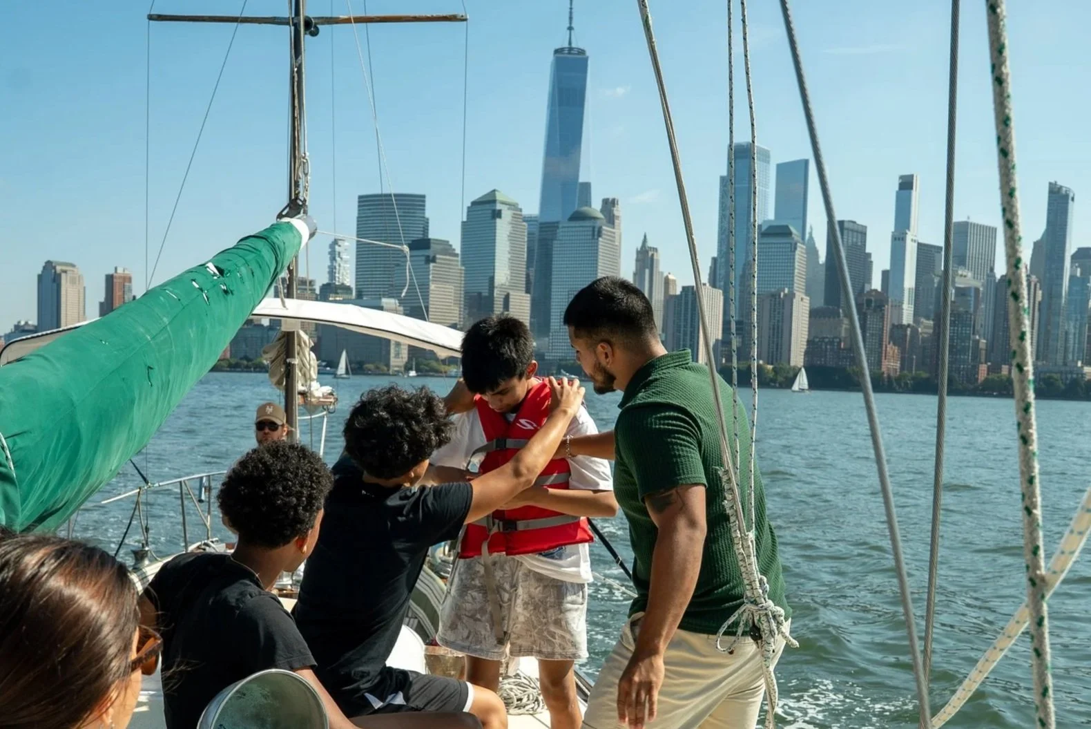 Group of people on a sailboat, with a city skyline including tall buildings and One World Trade Center in the background, during daytime.
