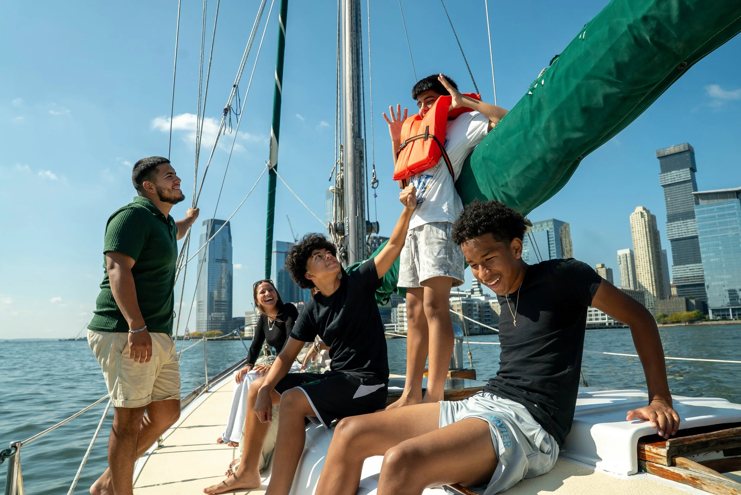 Young people enjoying a boat ride in a city harbor, with skyscrapers in the background, during daytime.