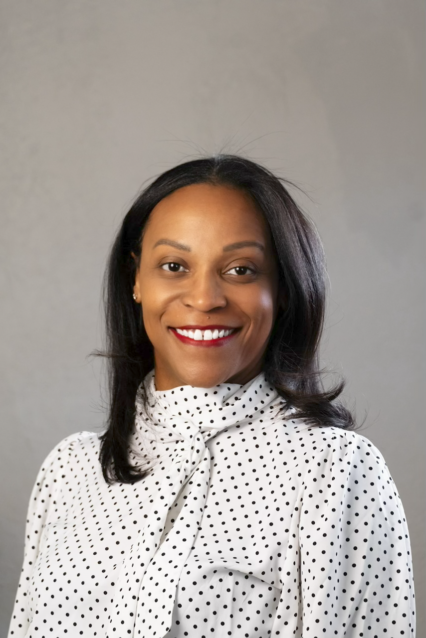 A portrait of a smiling African American woman with black hair, wearing a white blouse with black polka dots and a bow at the neck, against a plain gray background.