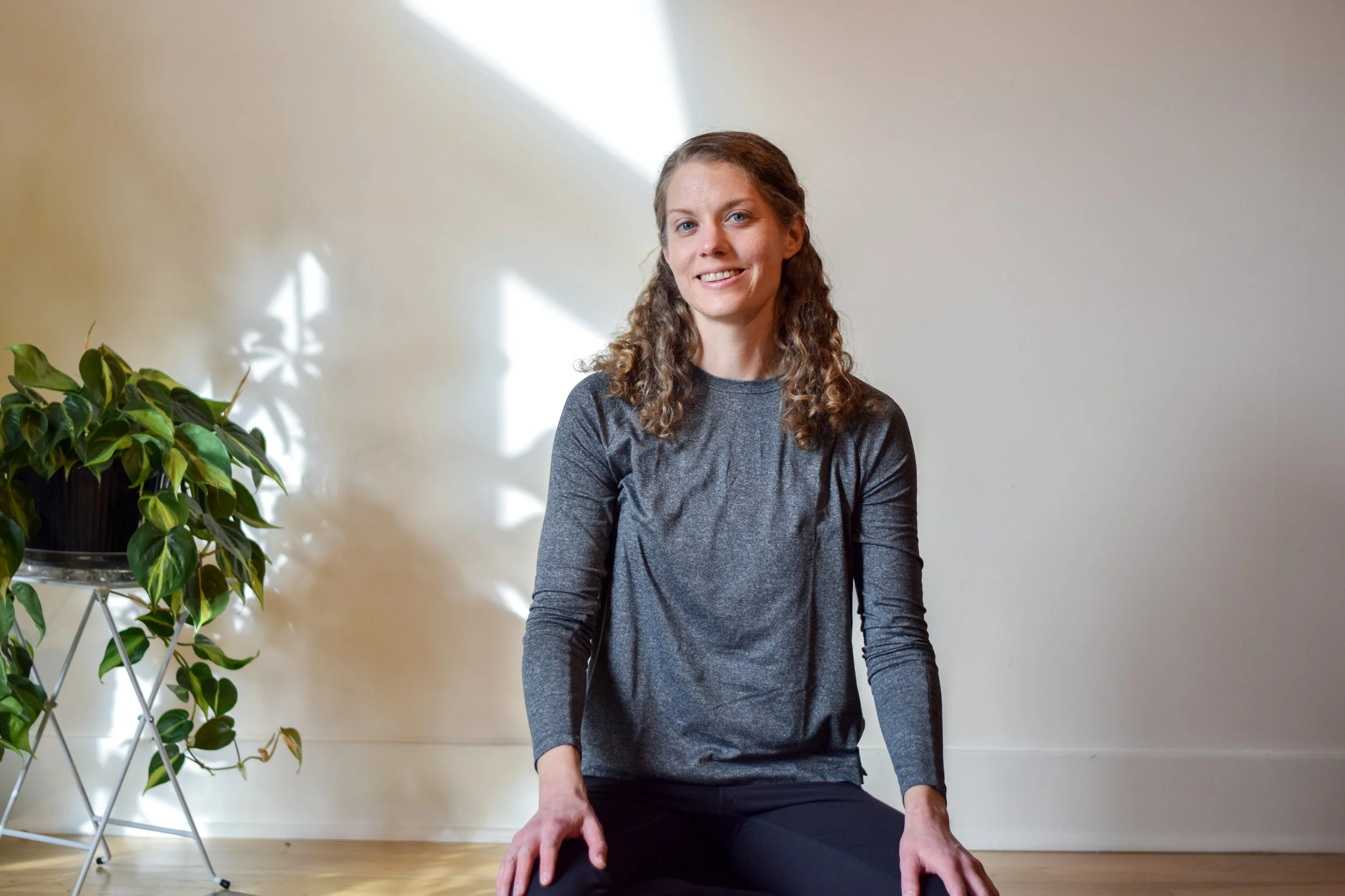A woman with curly blonde hair sitting on the floor near a potted plant, smiling in a room with sunlight and shadows on the wall.