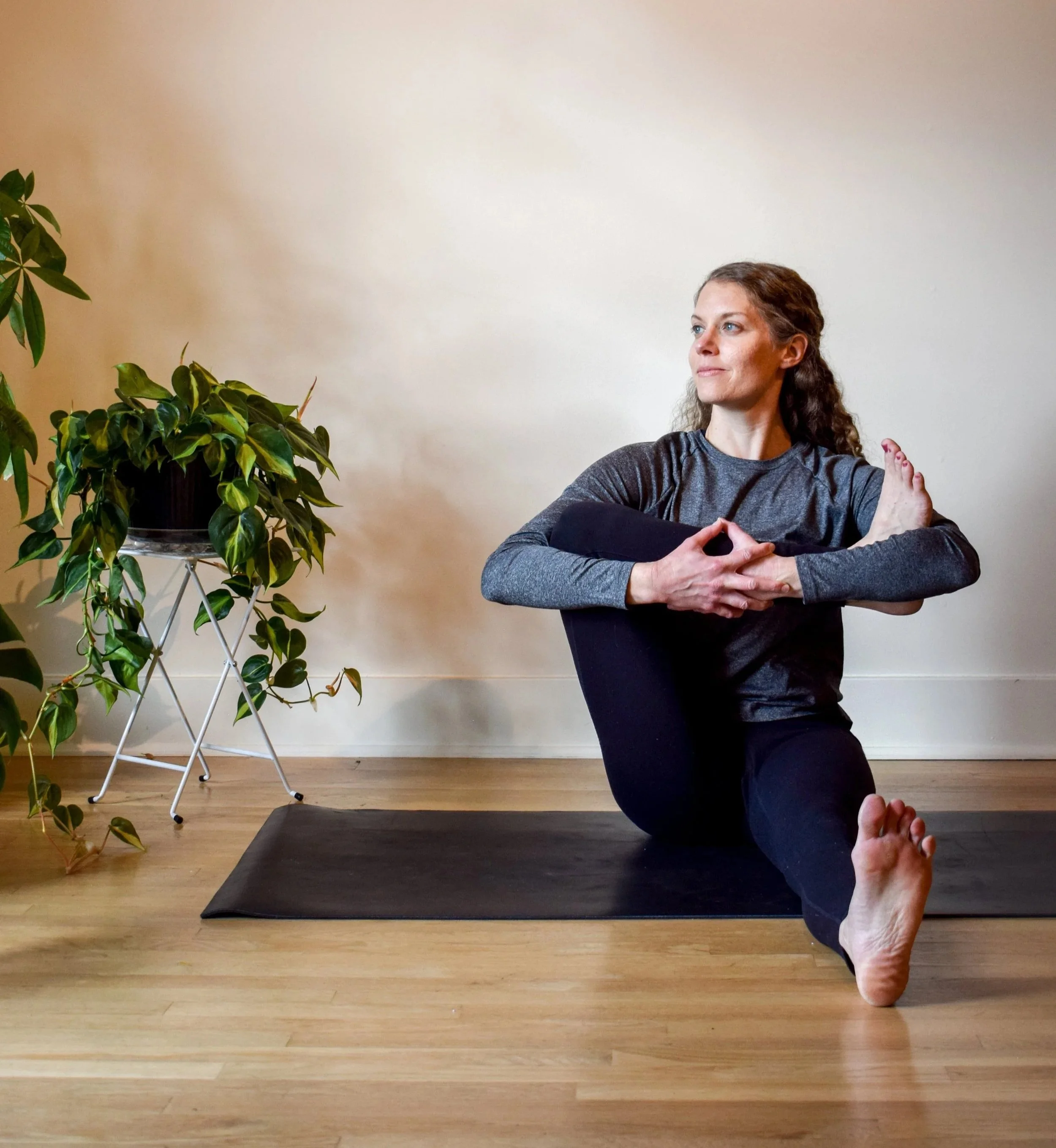 Woman doing yoga pose on black mat in a room with a plant on a stand and hardwood floor